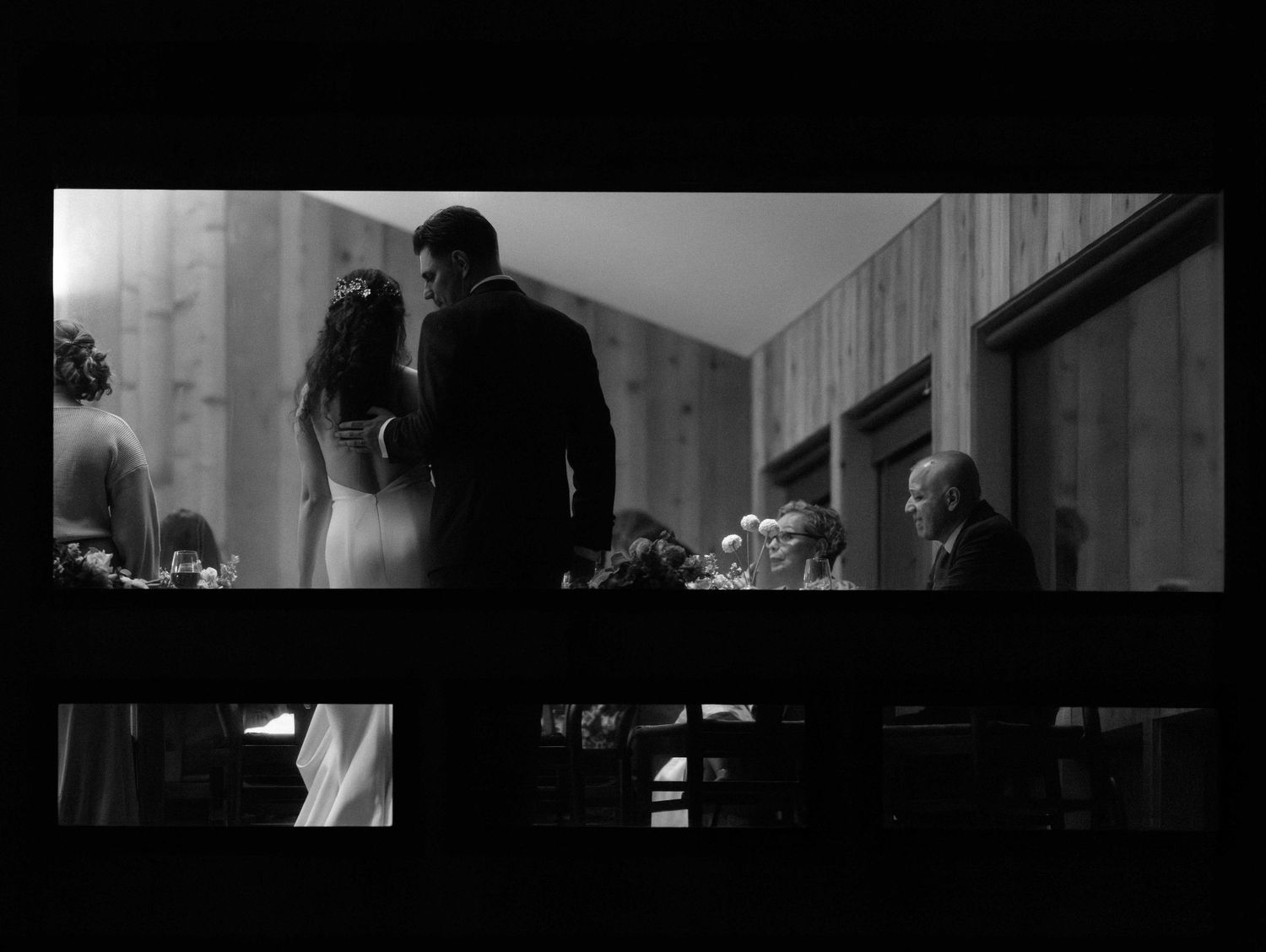 Black and white photo of groom gently touching bride on back as seen through a window.