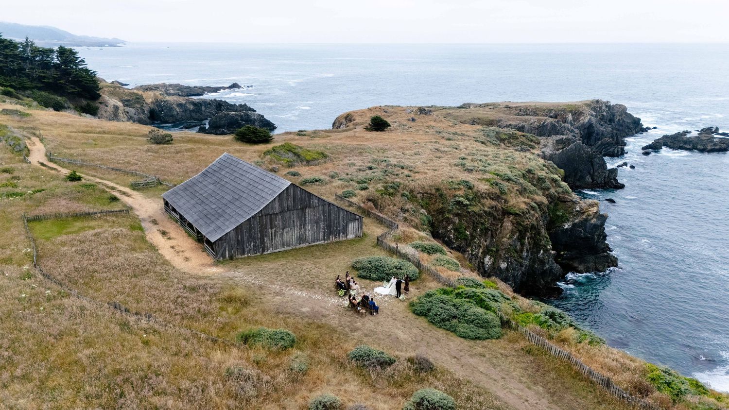 Aerial view of a rustic barn on coastal cliffs with horses grazing near the Pacific Ocean shoreline.