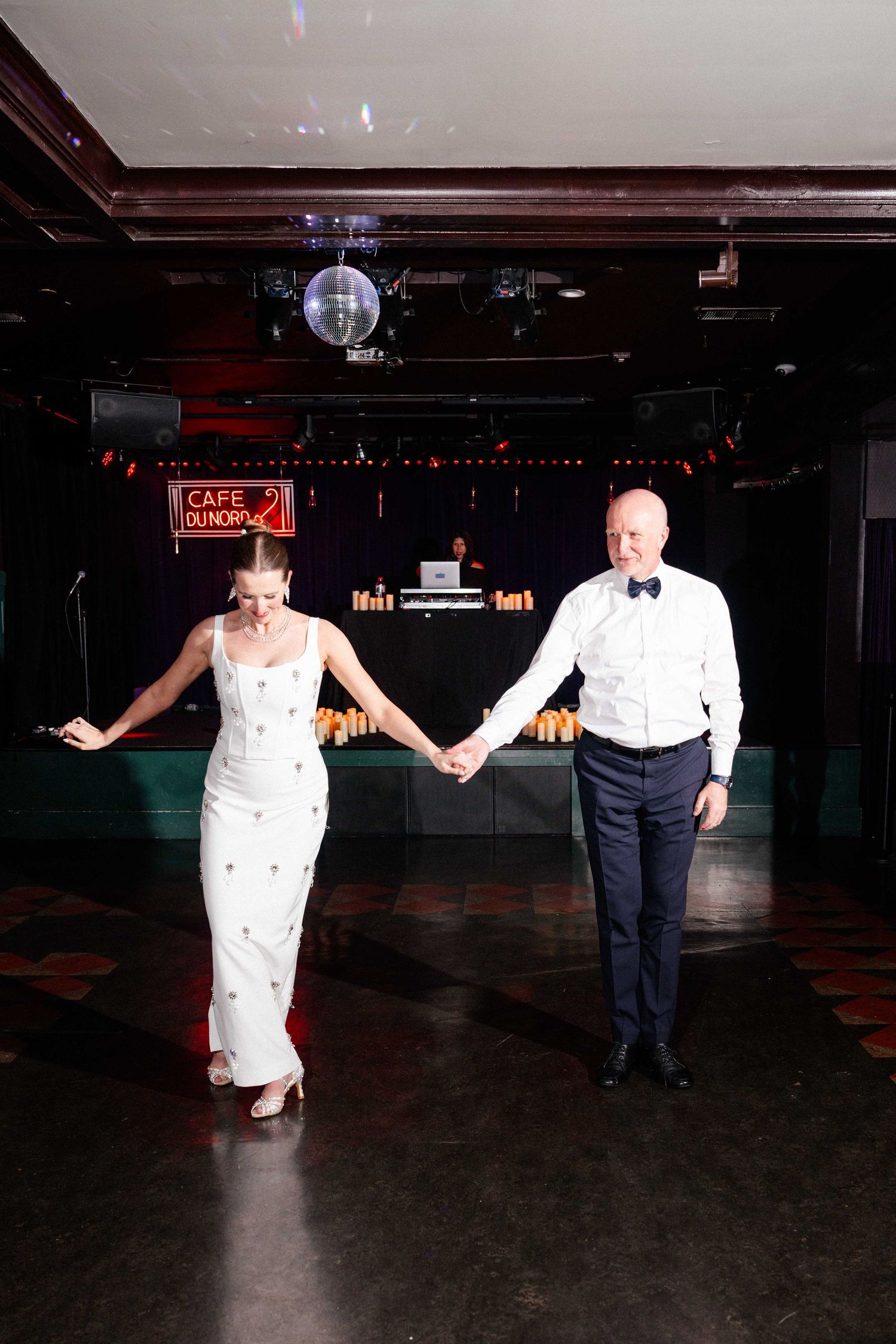 Bride and groom holding hands on dance floor at elegant wedding reception with disco ball and neon signs.