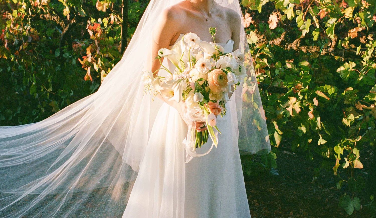 Bride in flowing white gown and veil holds elegant bouquet of white and peach flowers outdoors.