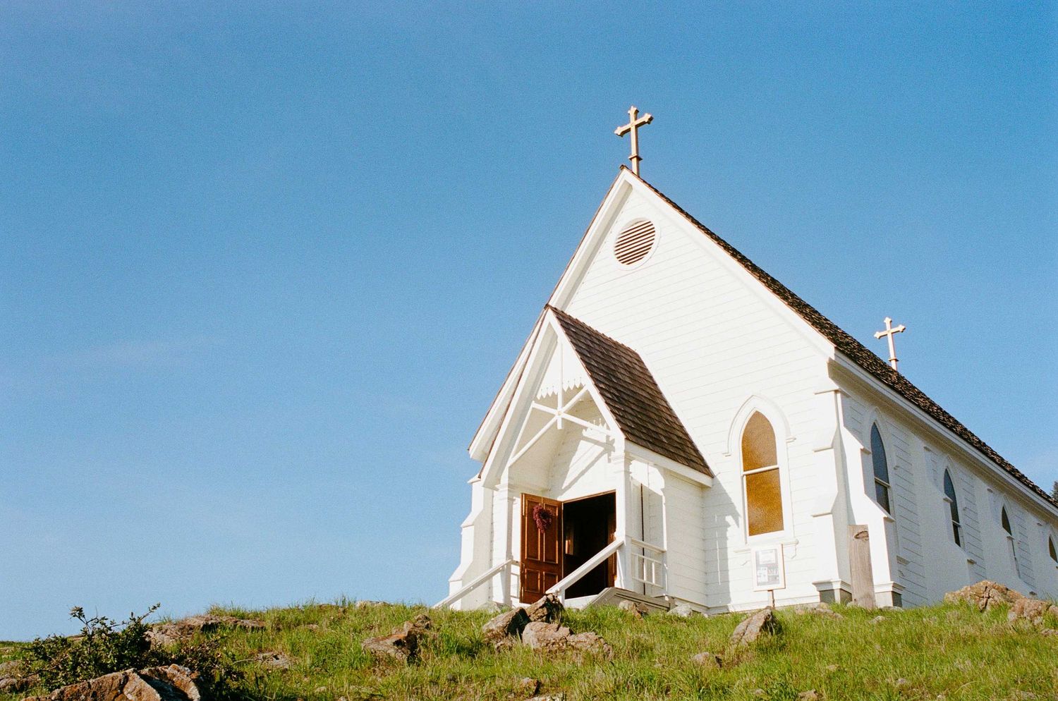 White wooden church with cross on steeple sits atop a grassy hill against a bright blue sky.