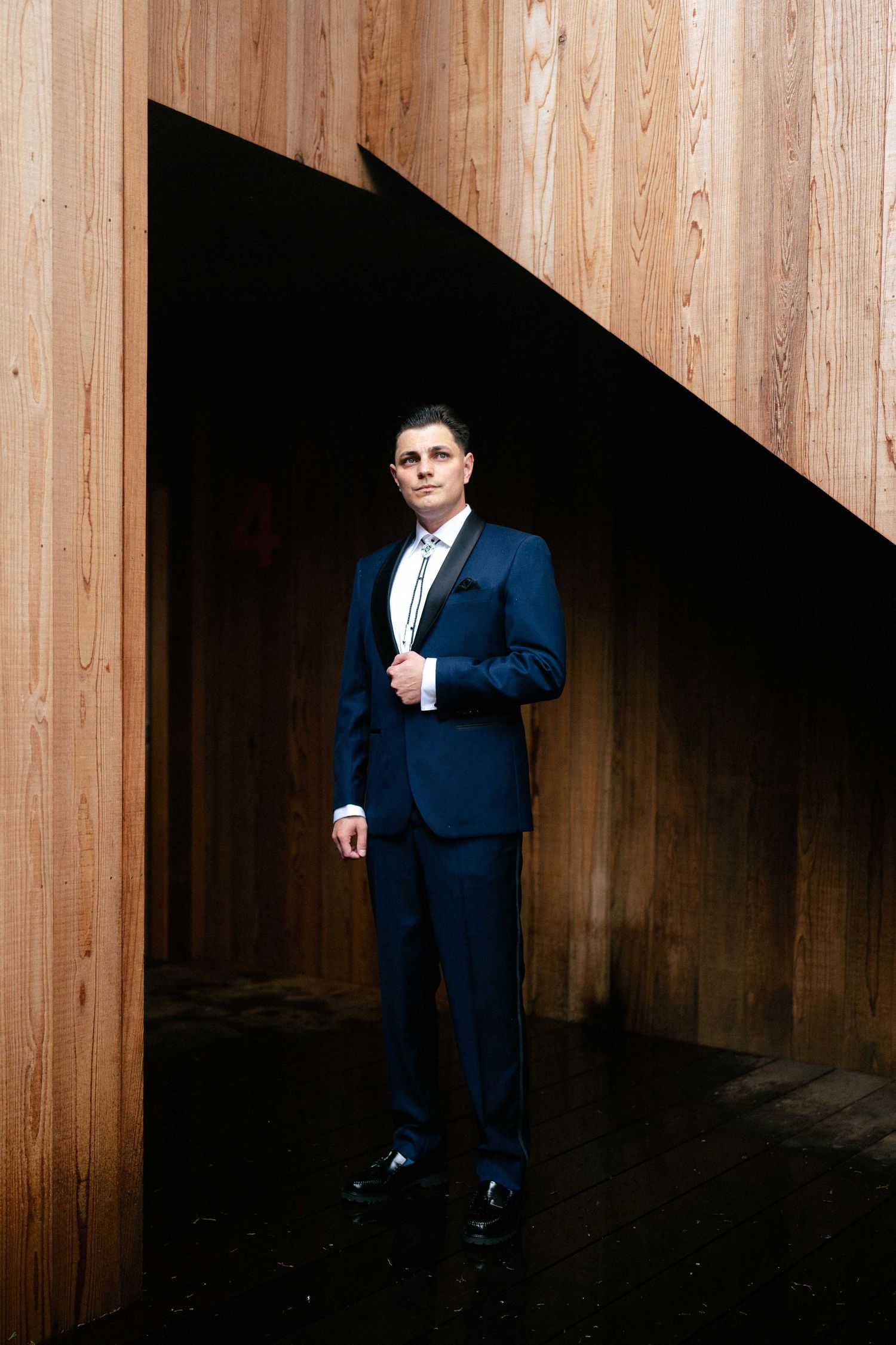 Man in navy blue suit standing between wooden panels in dramatic lighting.