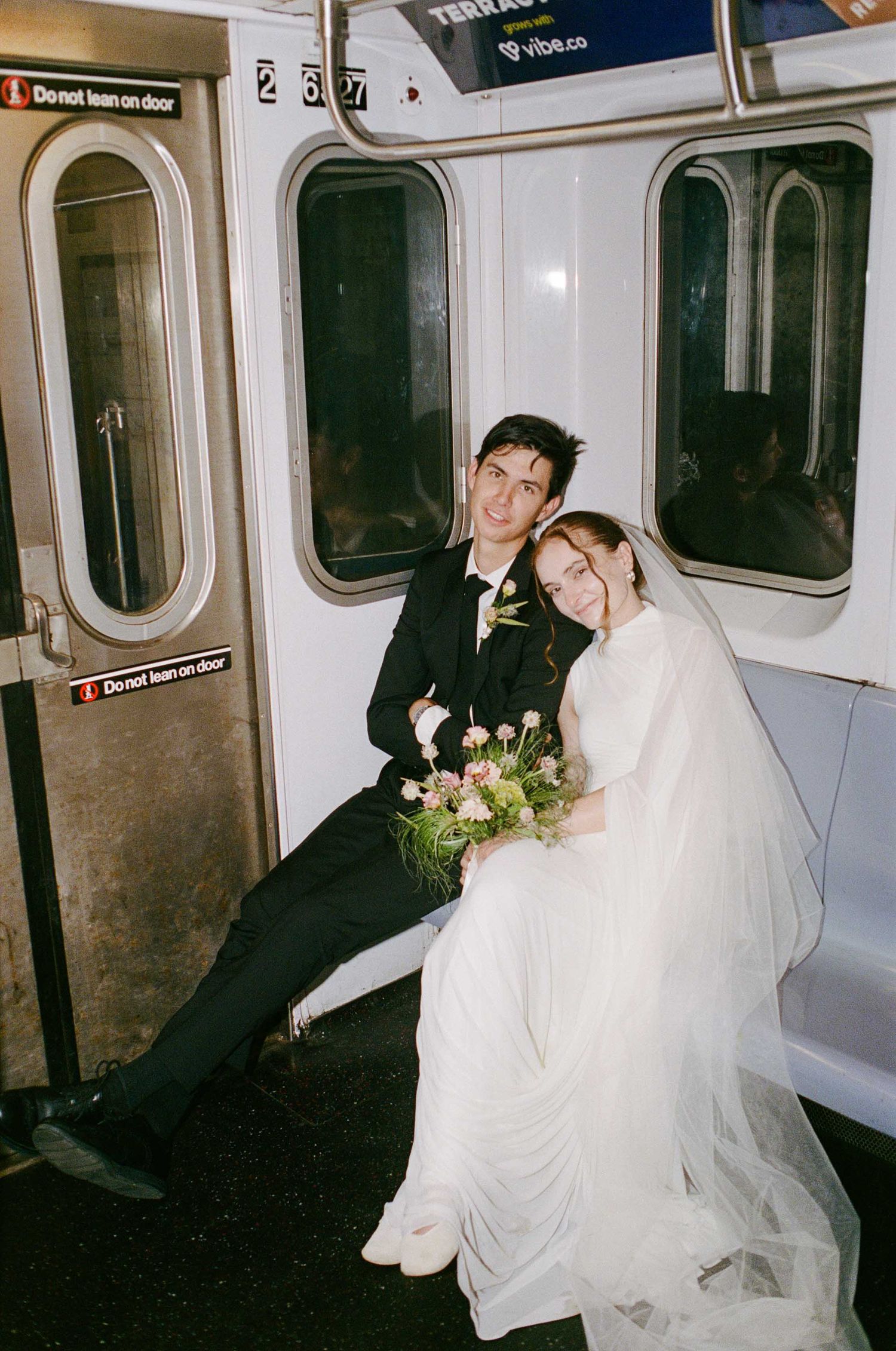 Newlywed couple in wedding attire sitting on a subway train, bride holding a floral bouquet, smiling together.
