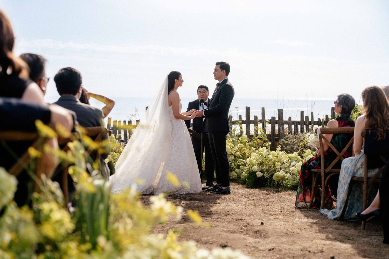 Outdoor wedding ceremony with bride and groom exchanging vows near a rustic fence, surrounded by yellow flowers and ocean views.