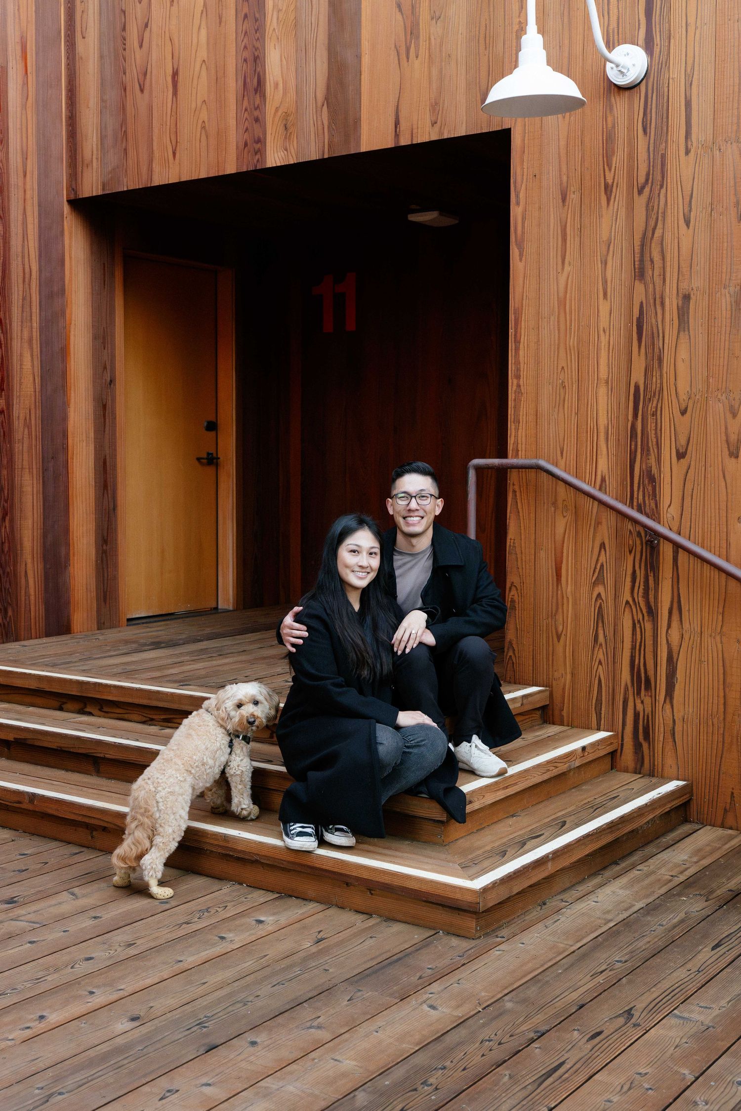 A couple and their fluffy dog sit on wooden steps outside a modern cedar-clad building entrance.