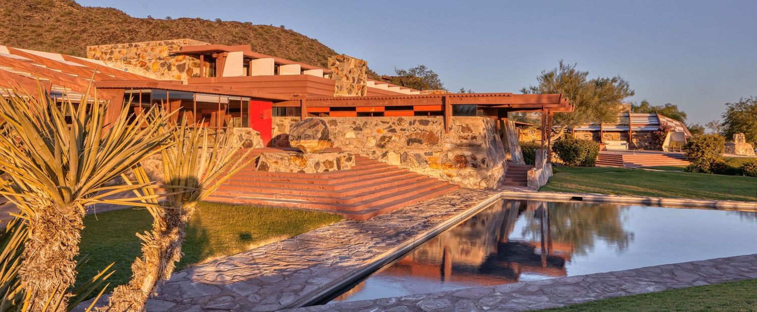 Frank Lloyd Wright-inspired desert estate with red stone architecture, reflecting pool, and desert landscaping at sunset.