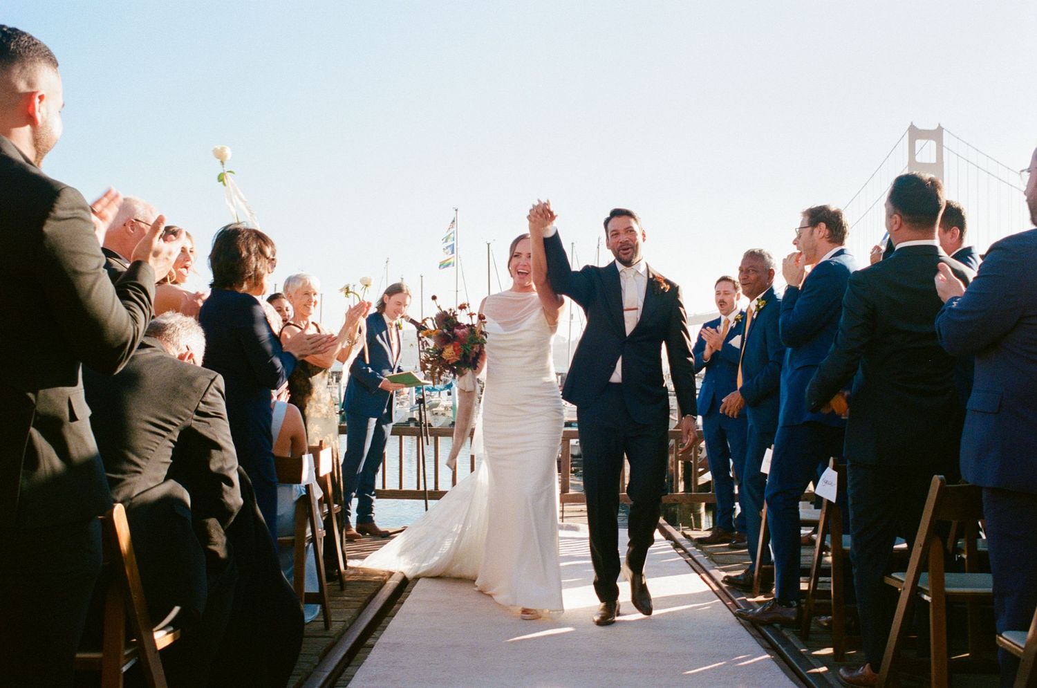 Newlyweds walk triumphantly down aisle as guests cheer with Golden Gate Bridge visible behind.