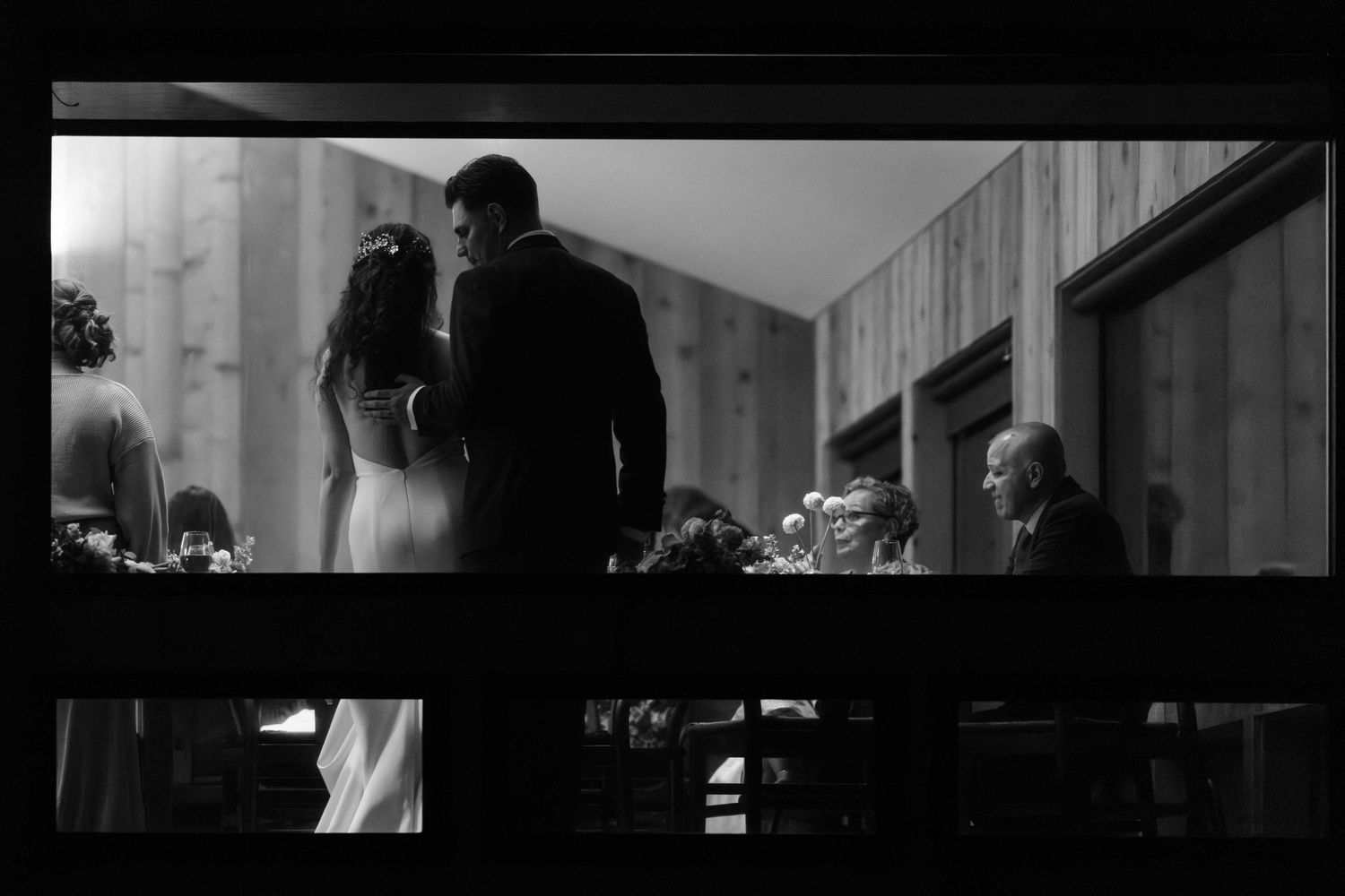 Black and white photo of groom gently touching bride on back as seen through a window.