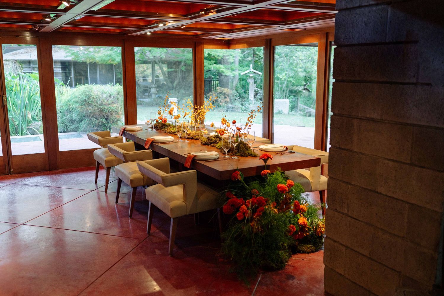 A long wooden wedding reception table set with floral centerpieces in a sunlit room with garden views.