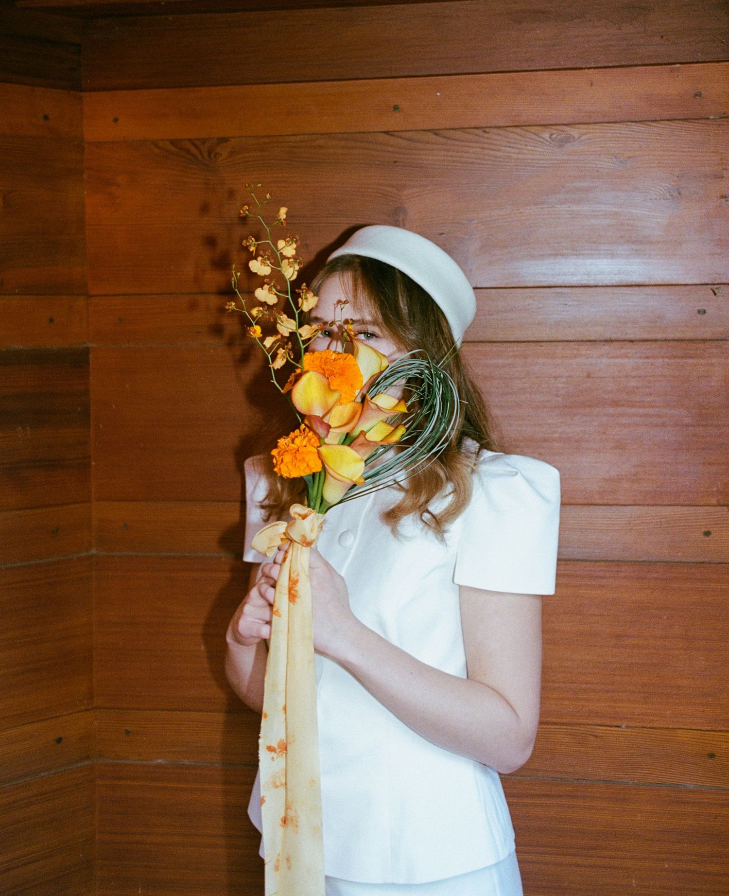 Bride in white mod dress and pillbox hat holding a vibrant orange and yellow bouquet against a wood-paneled wall.