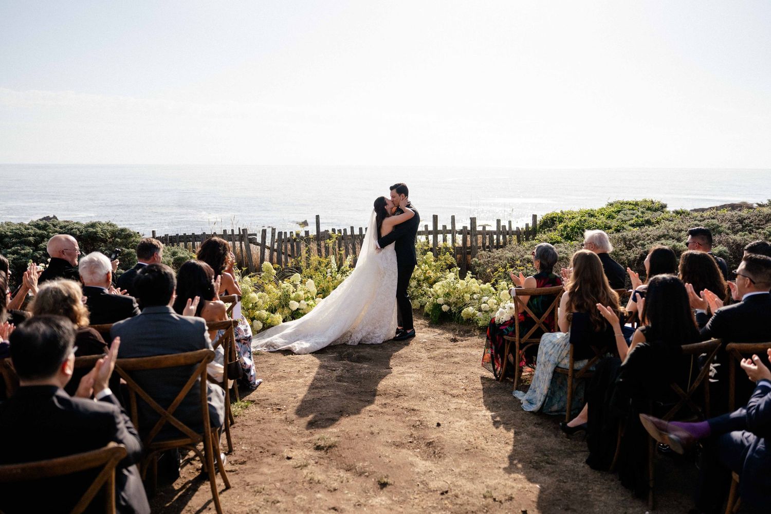 Bride and groom share first kiss at outdoor cliffside wedding ceremony overlooking the ocean with seated guests.