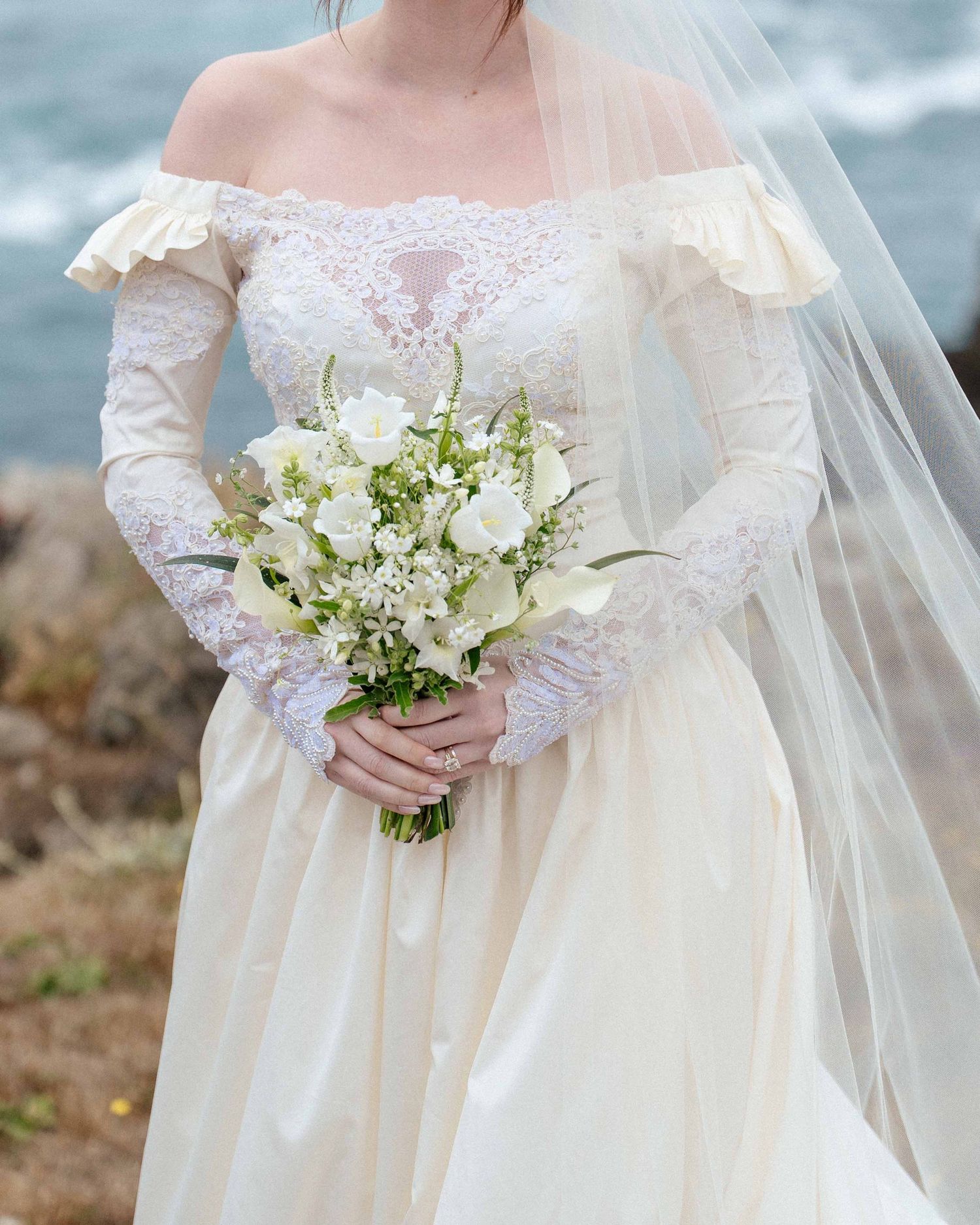 Bride in off-shoulder lace wedding gown holding white floral bouquet with veil, standing near ocean cliffs.