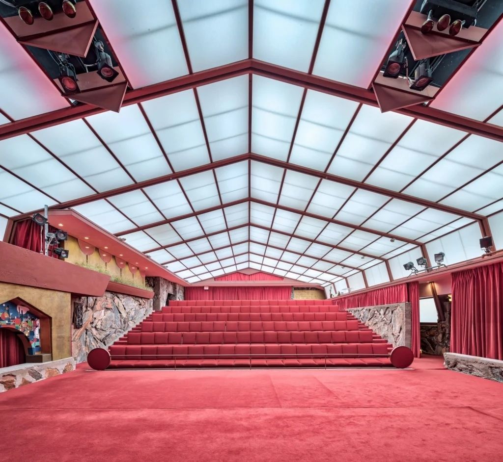 Dramatic red theater interior with tiered seating, translucent ceiling panels, and stage lighting in a mid-century modern auditorium.