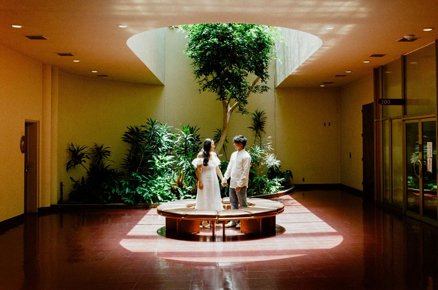 Couple standing on a circular platform surrounded by lush indoor plants under a skylight in an elegant atrium.