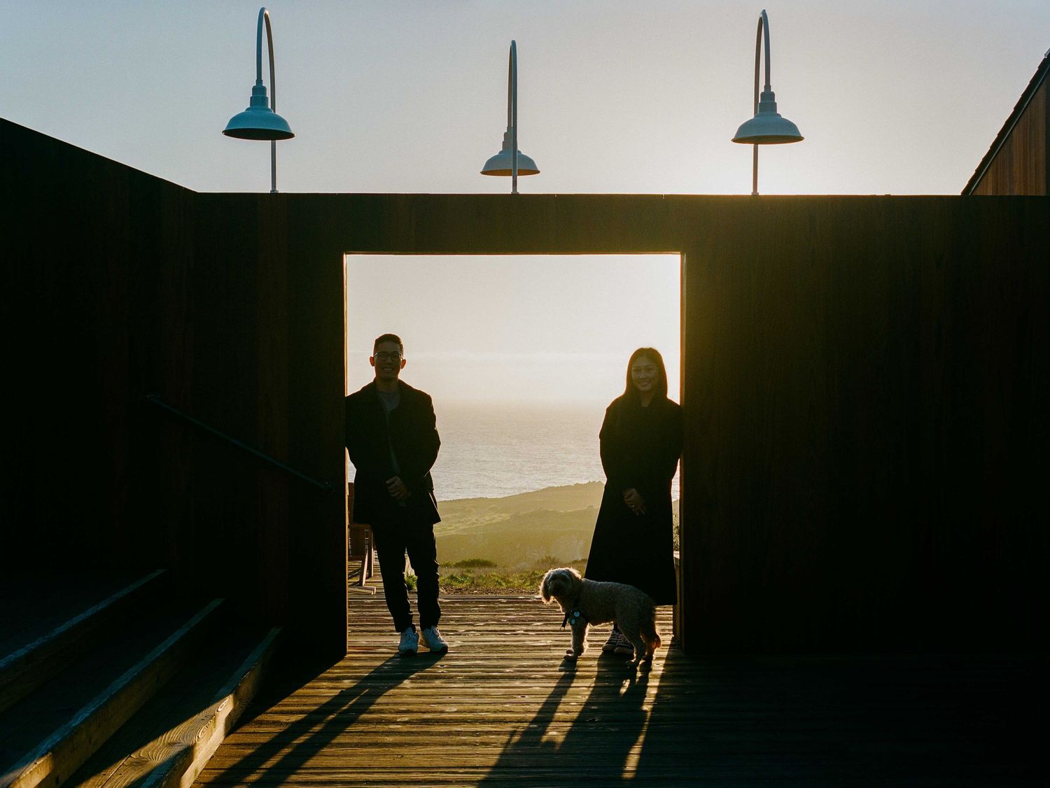Two silhouetted figures and a dog stand in a wooden gateway framing a stunning ocean sunset view.