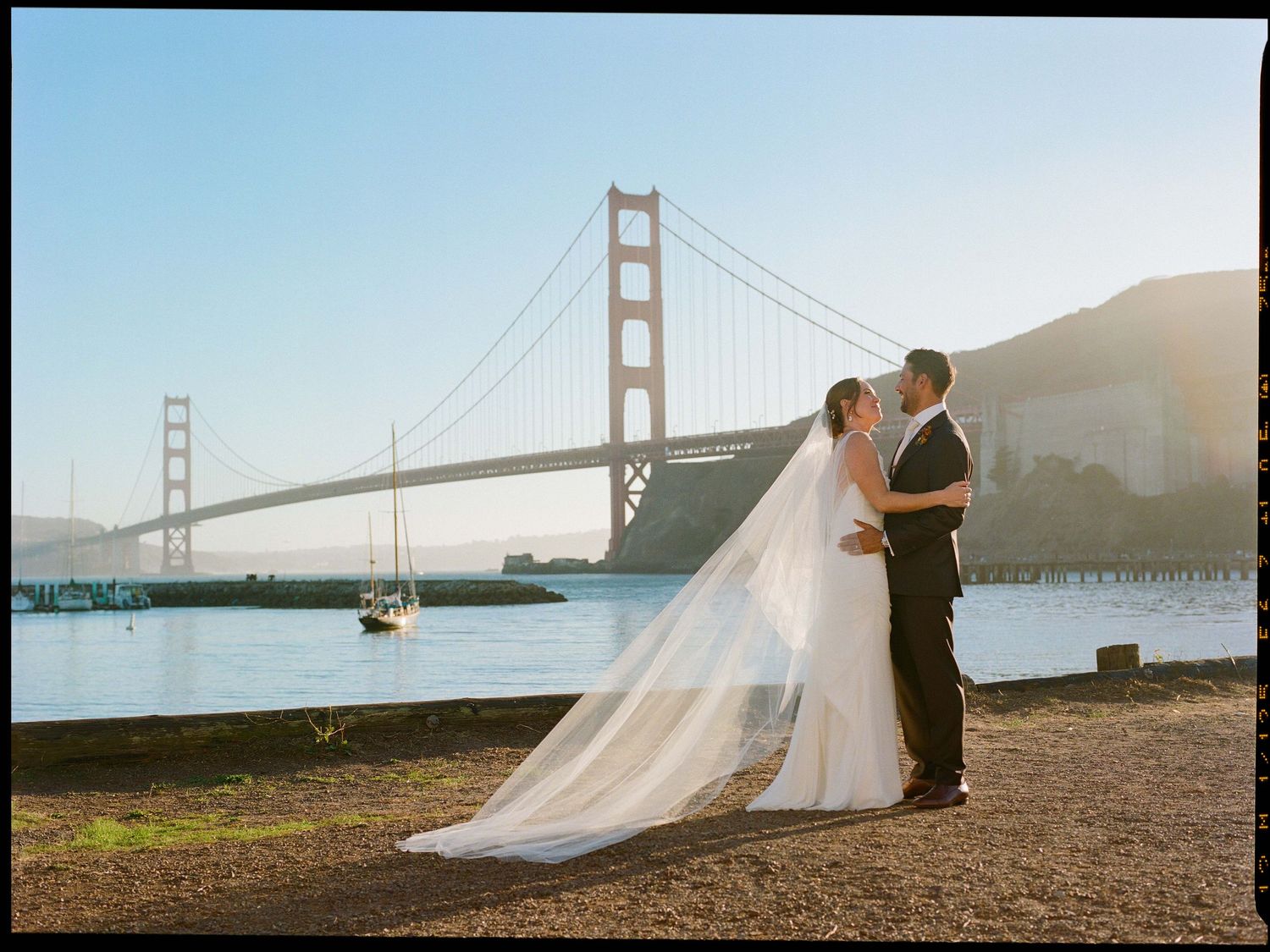 Bride and groom embrace near Golden Gate Bridge on sunny wedding day in San Francisco.
