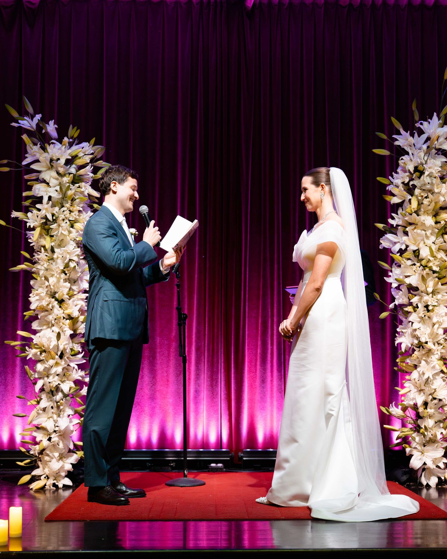 Bride and groom exchange vows at elegant wedding ceremony with white floral columns and purple draped backdrop.