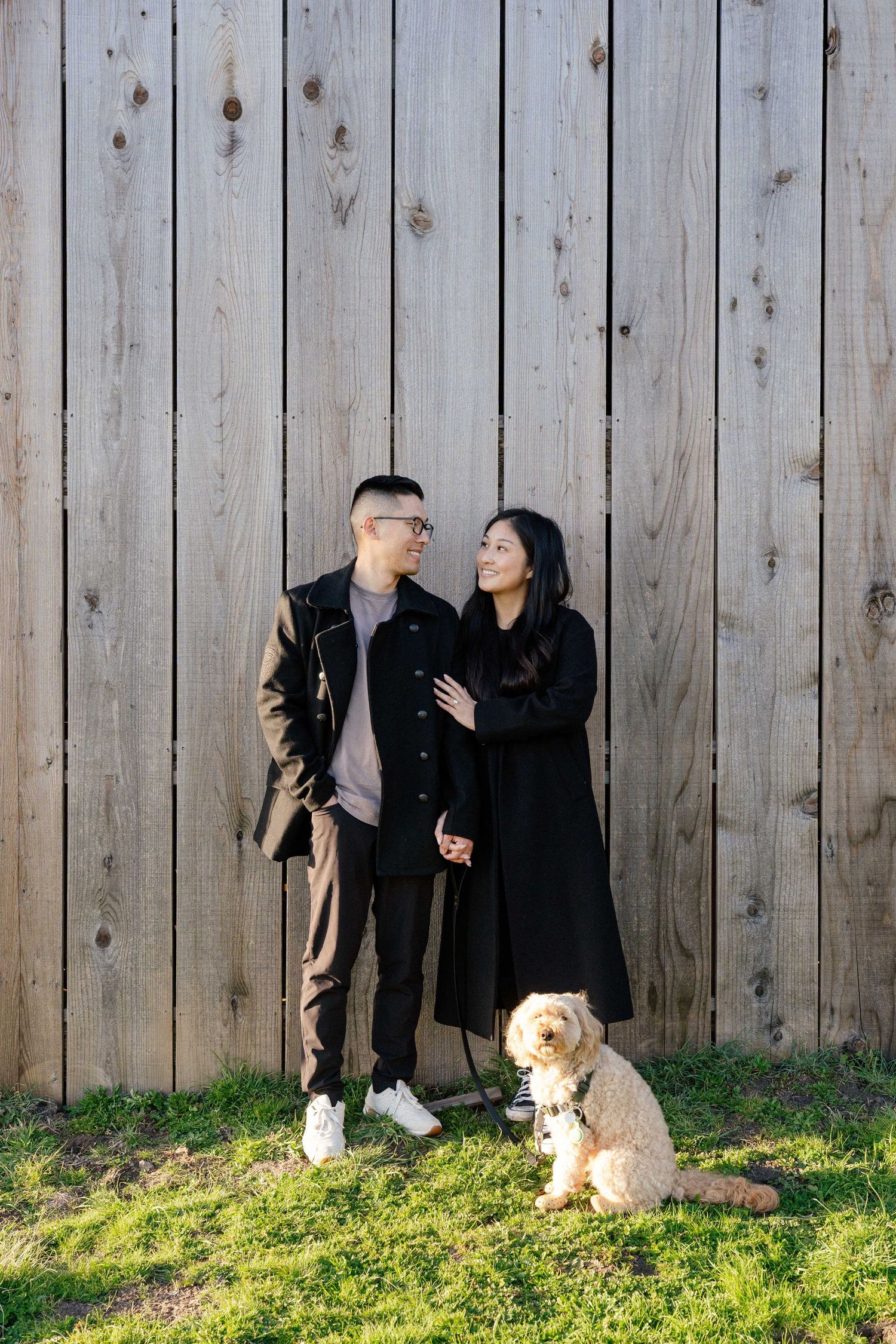 Couple holding hands with a fluffy dog, standing against a wooden fence on green grass.