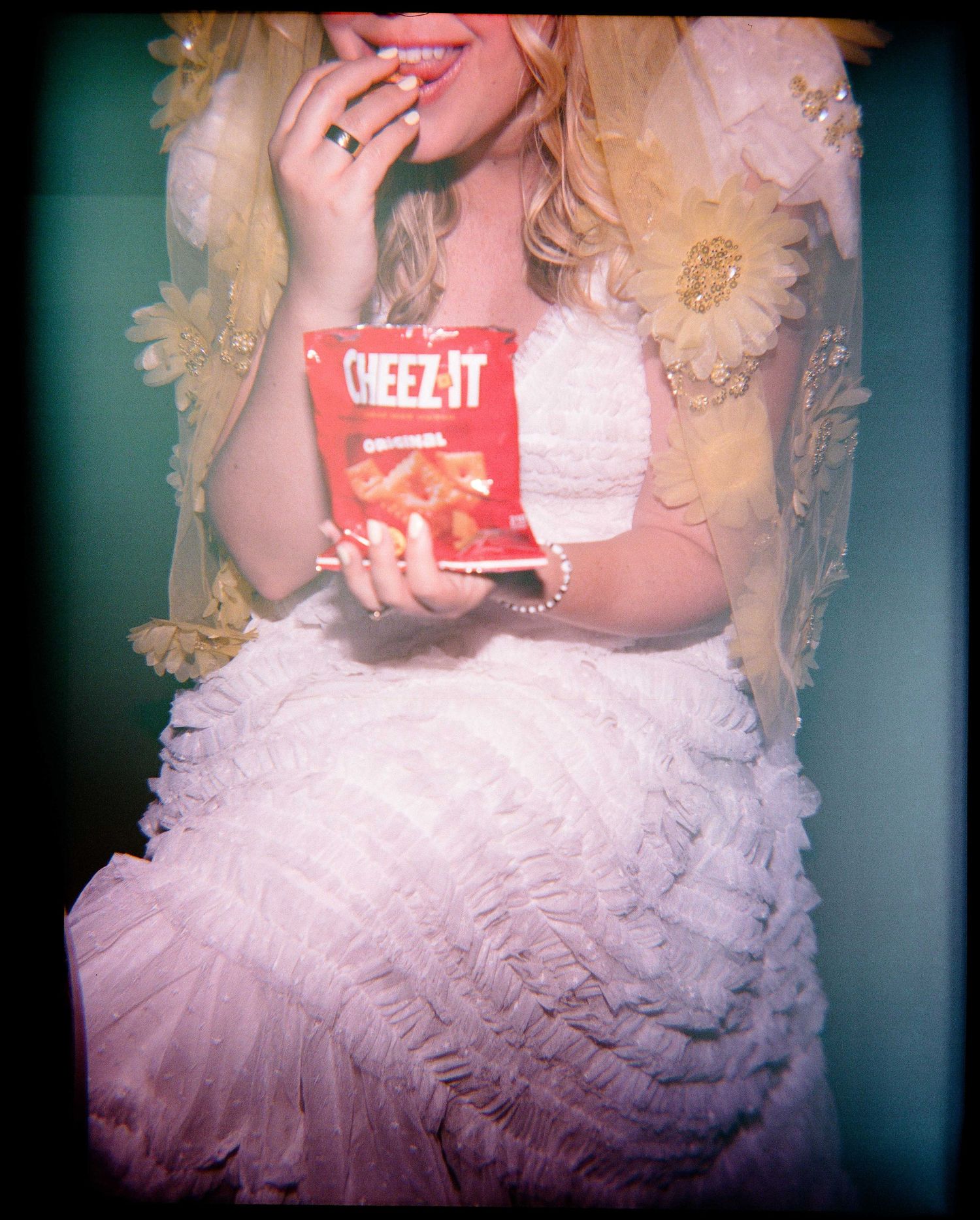 Woman in white ruffled dress holding Cheez-It box, wearing pearl bracelet and ring, eating crackers.