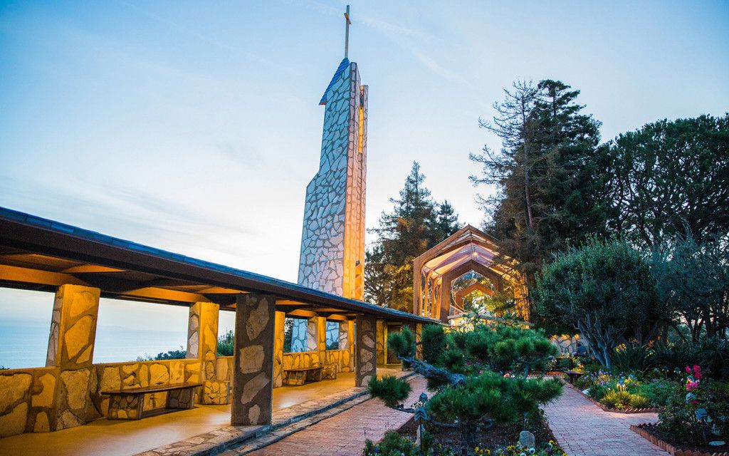 Dramatic stone chapel tower with cross at dusk, surrounded by lush gardens and rustic architecture.