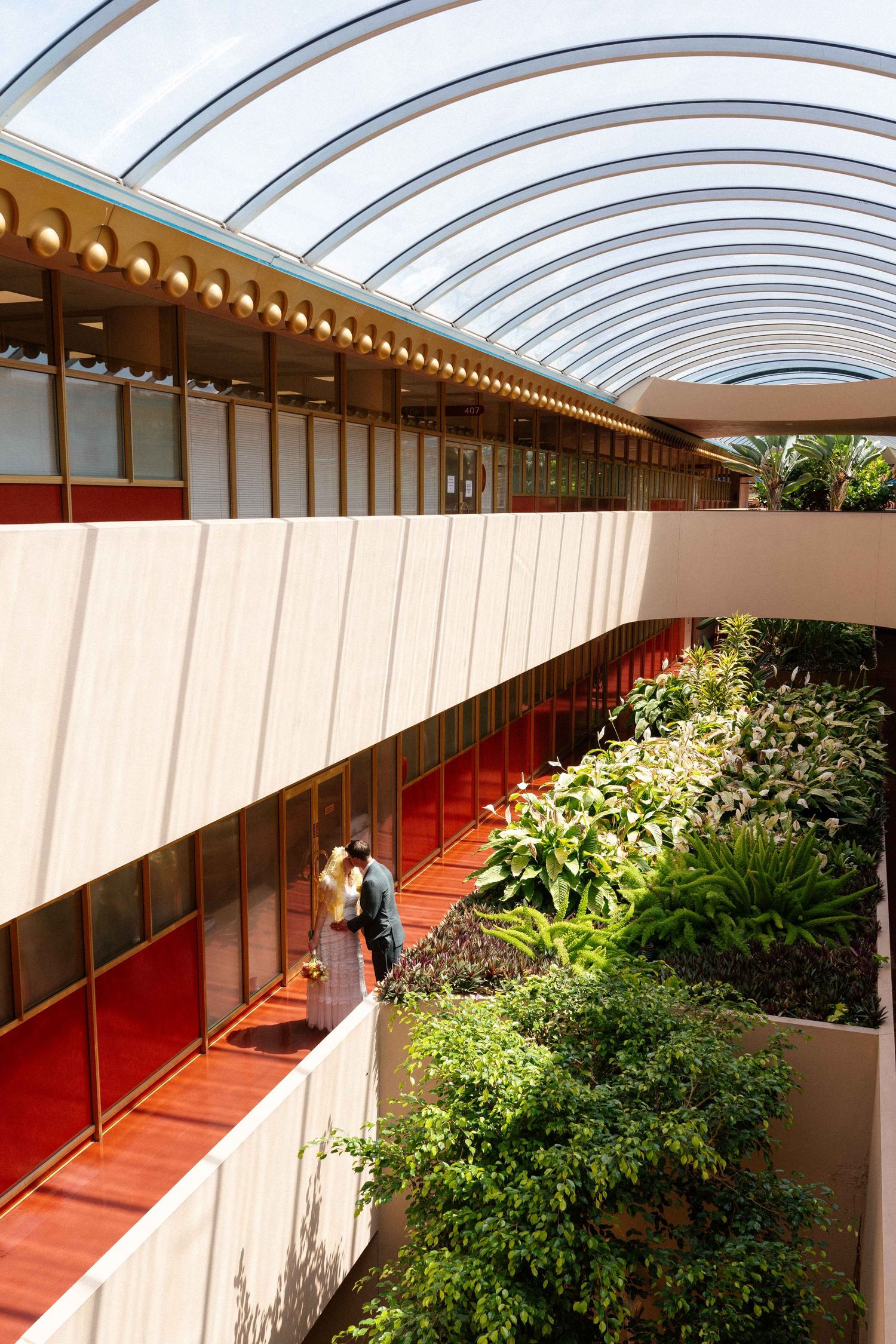 Couple walks along a curved indoor atrium corridor with lush greenery and arched glass ceiling.