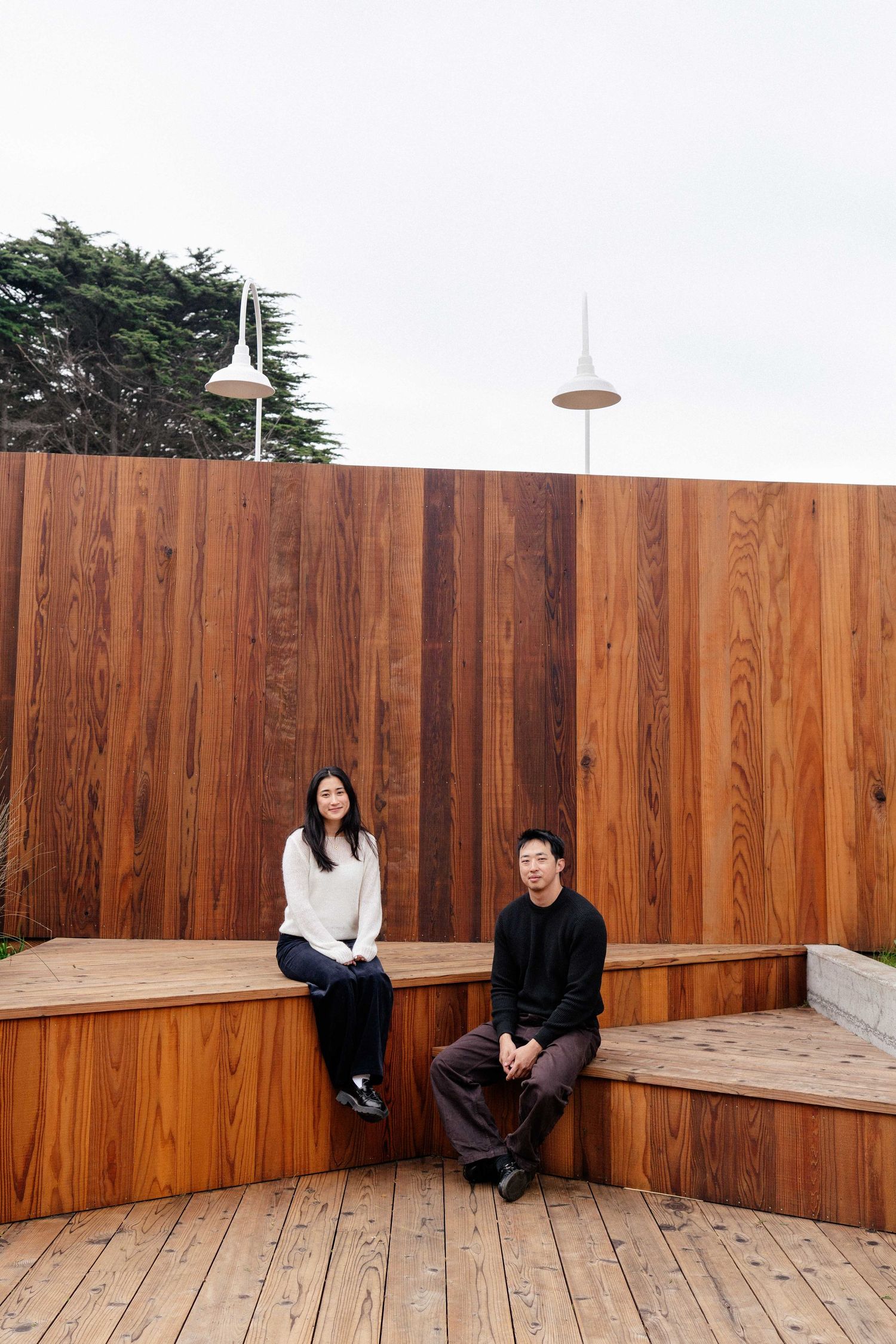 Two people sitting on a wooden bench against a tall cedar wood panel wall on a deck outdoors.