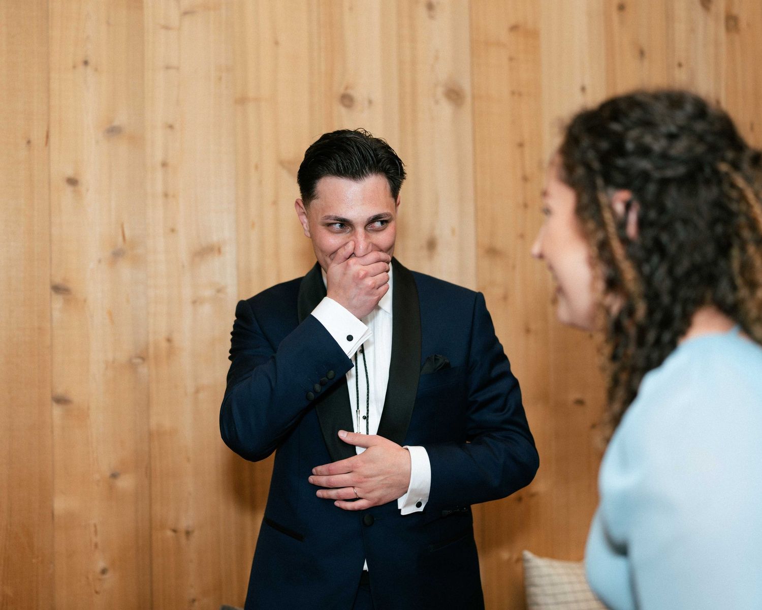 Groom in black suit covers mouth in emotional reaction during wedding first look moment indoors.