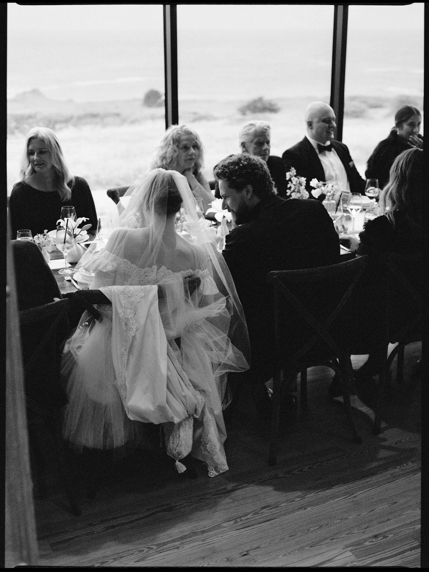 Black and white wedding reception photo of bride in veil and gown seated at elegant dinner table with guests.