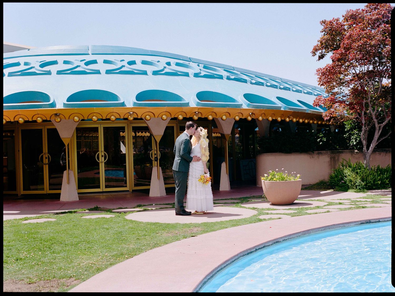 Bride and groom share a romantic moment outside a colorful domed wedding venue beside a pool.