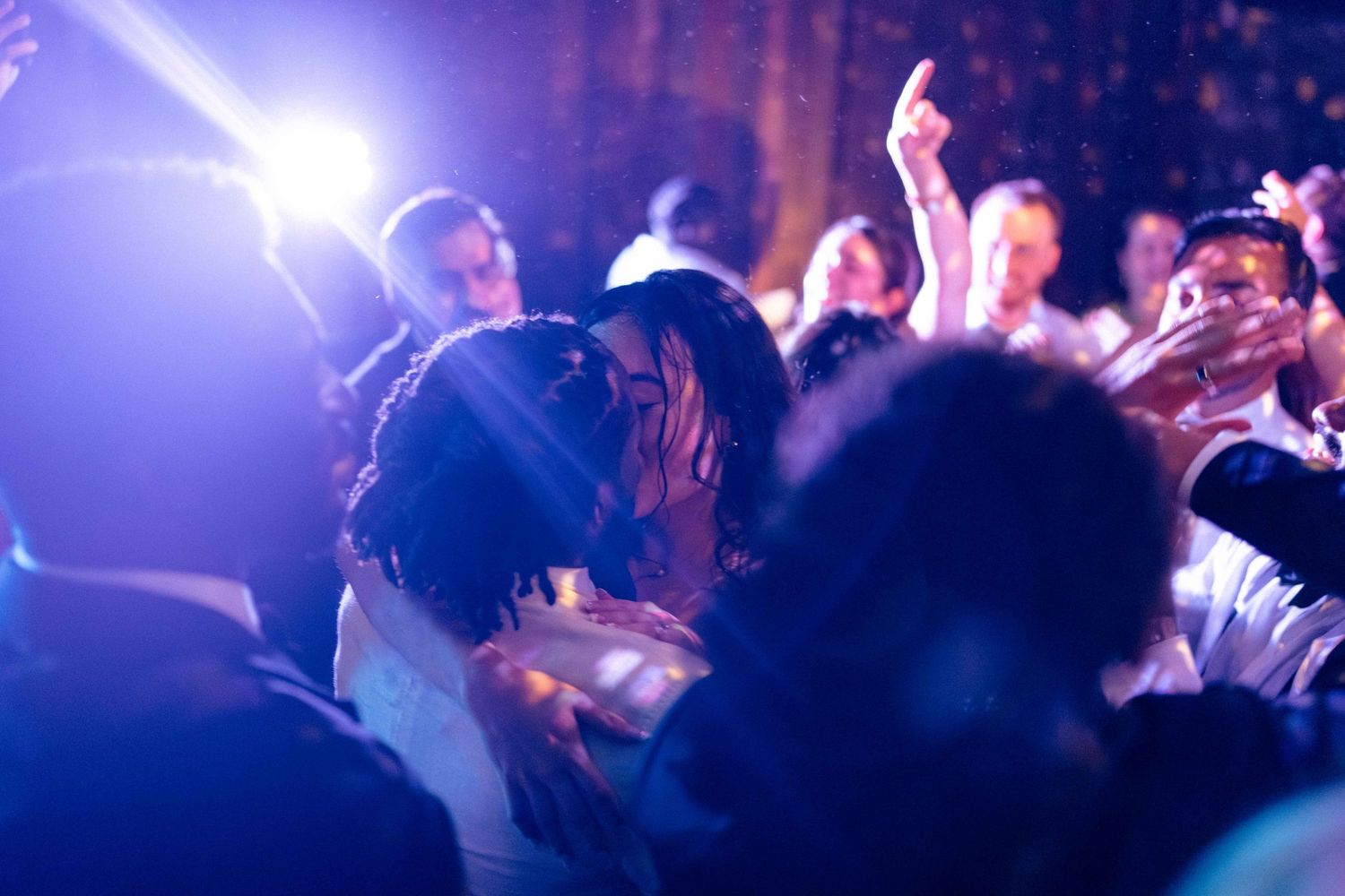 Couple sharing a romantic kiss on the dance floor surrounded by wedding guests in soft blue lighting.