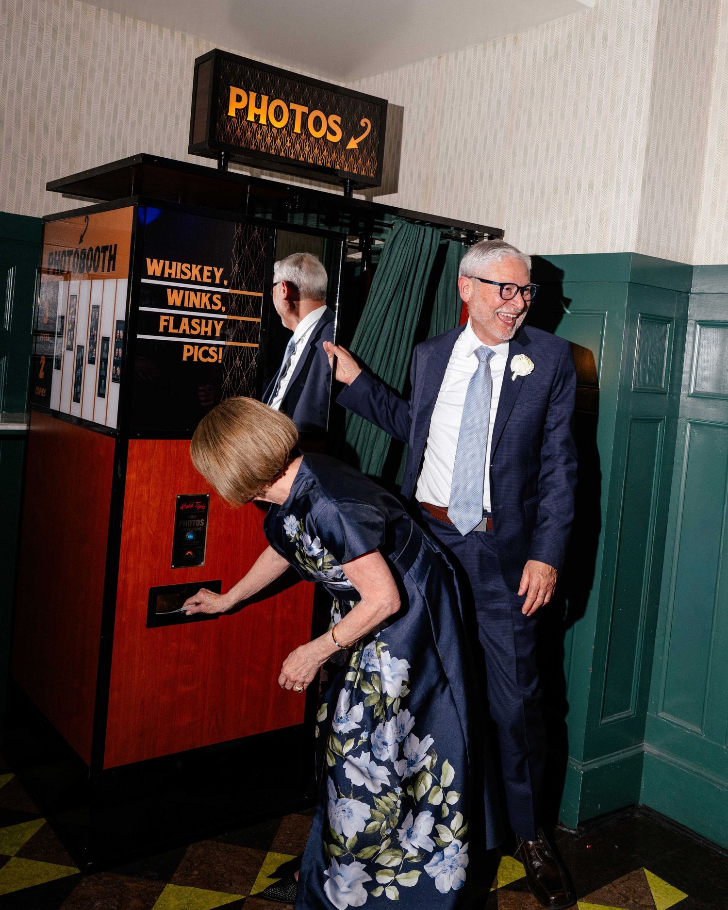 Guests enjoying a vintage photo booth at a wedding reception with a sign reading Whiskey, Winks, Flashy Pics.
