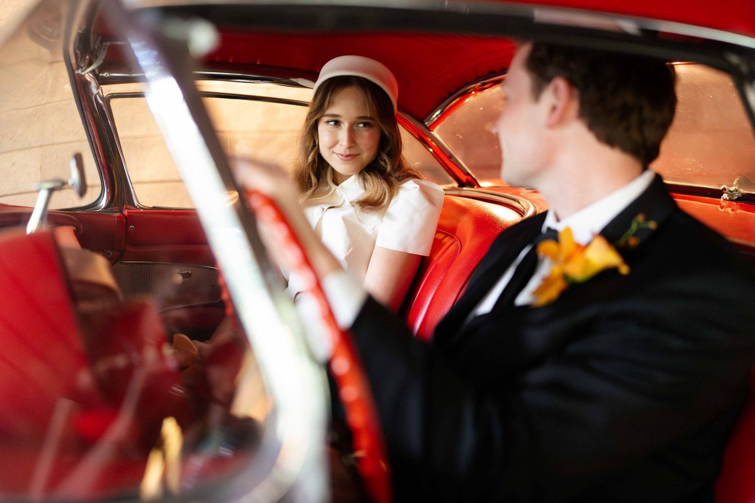 Bride in white dress and pillbox hat sitting with groom in a vintage red car interior during wedding day.