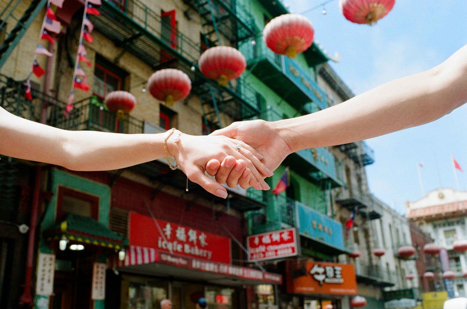 Two hands clasped together in San Francisco Chinatown with red lanterns and storefronts in the background.