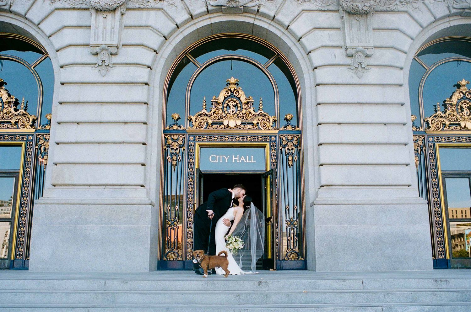 Bride and groom kiss on steps of San Francisco City Hall beneath ornate golden archway.