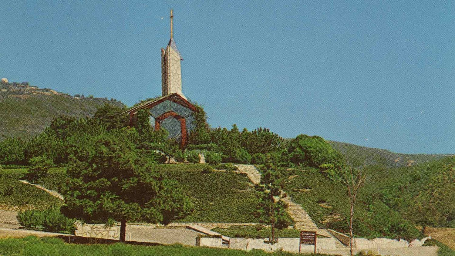 Small hillside chapel with tall steeple and cross, surrounded by terraced gardens and mountain backdrop.