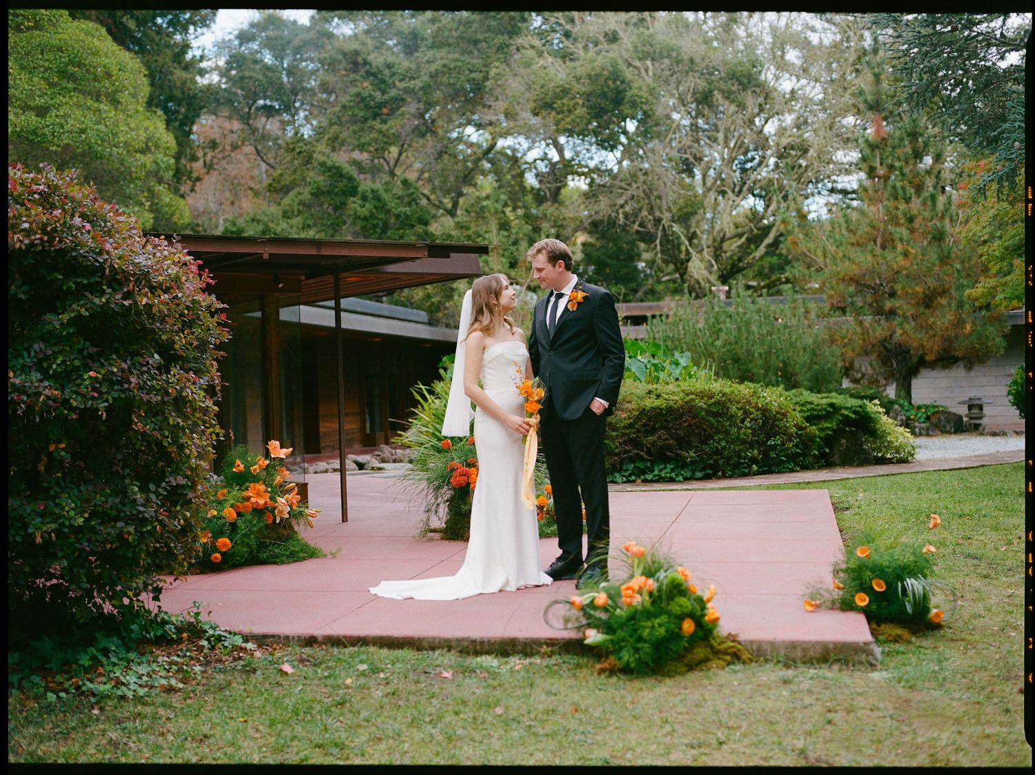 Bride and groom share intimate moment on terrace with orange floral arrangements nearby.