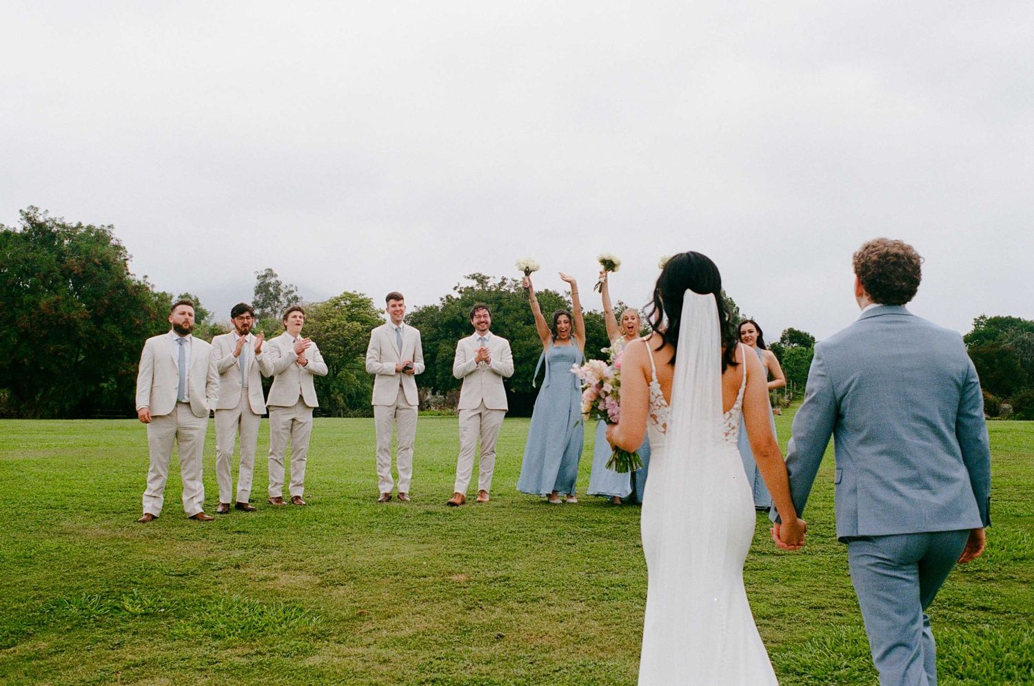 Wedding party cheers as bride and groom walk hand in hand across green lawn.