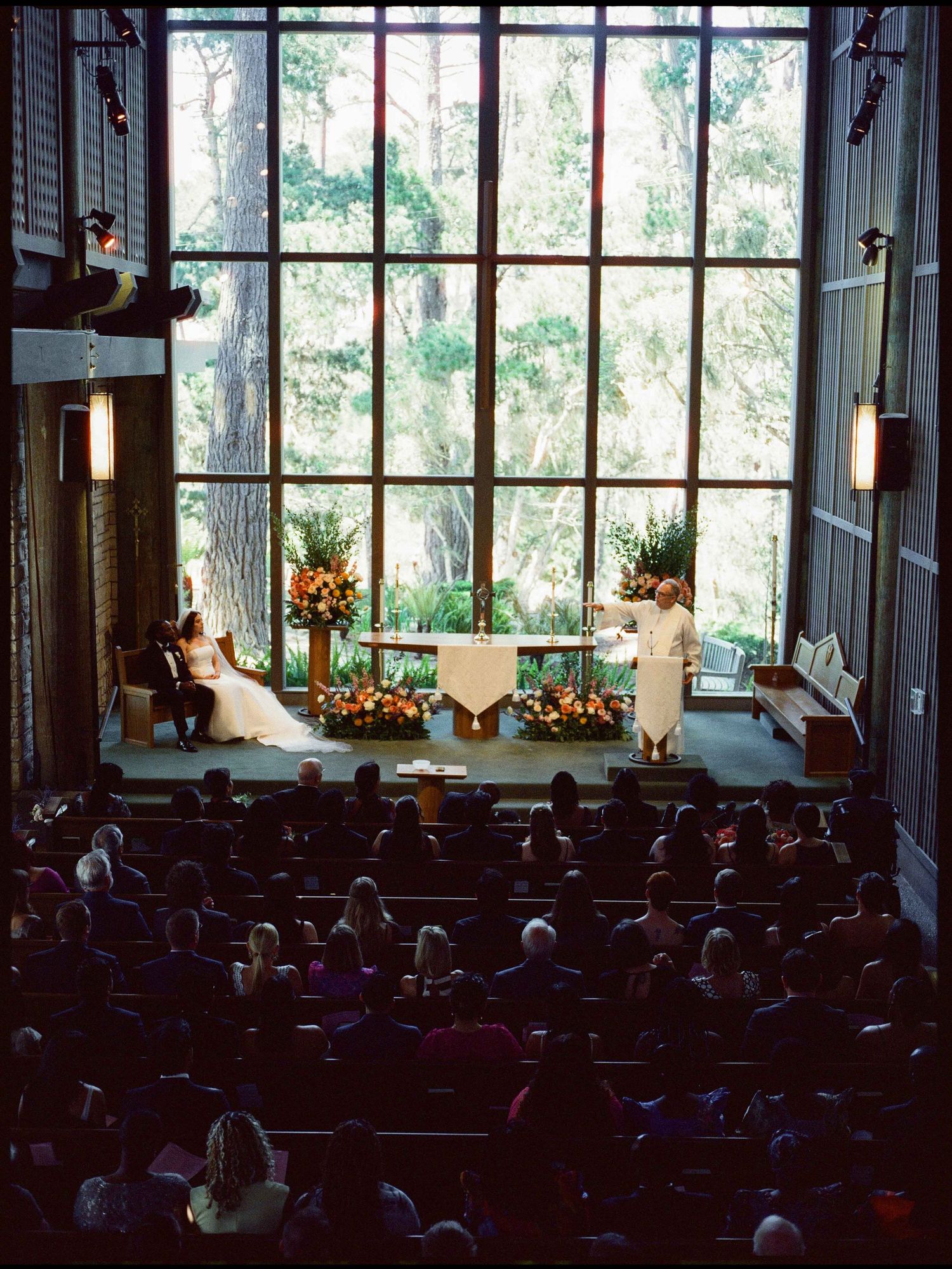 Elegant church wedding ceremony with guests seated in pews, couple at altar before large floor-to-ceiling windows overlooking trees.