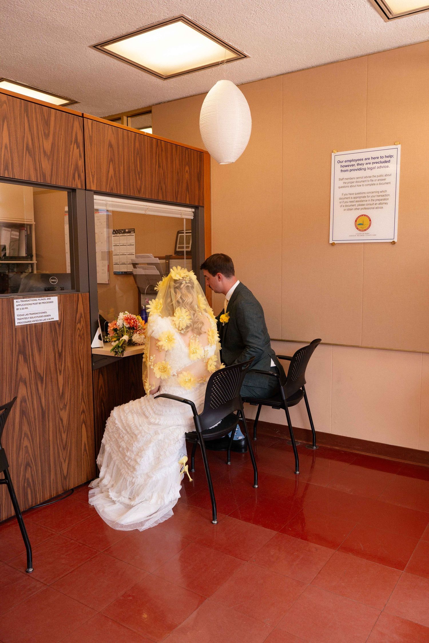 Bride and groom in wedding attire signing documents at a government office counter on their wedding day.