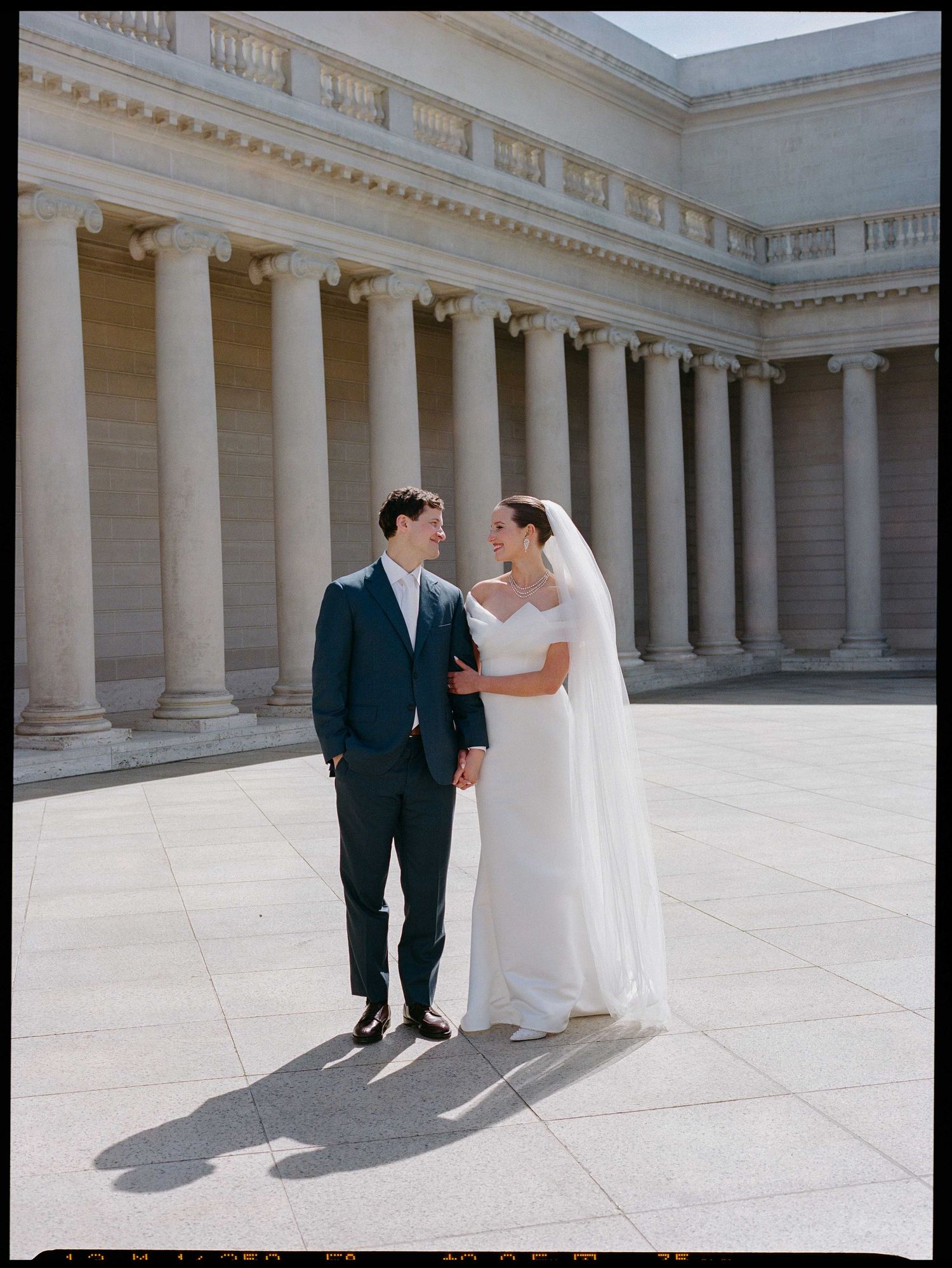Bride and groom share a romantic moment at a grand neoclassical building with tall columns on their wedding day.