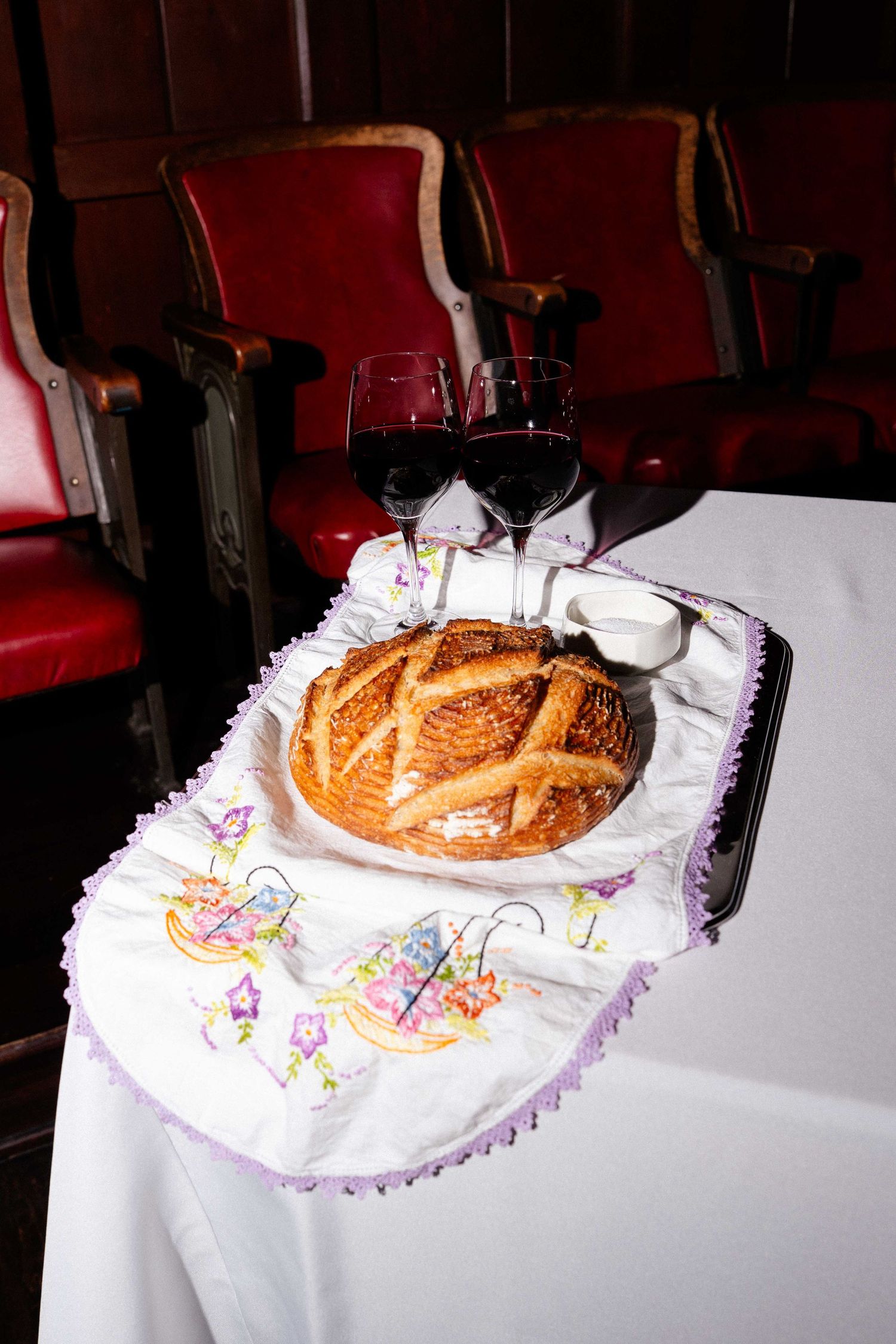 Traditional challah bread on embroidered cloth with two glasses of red wine on a white table.