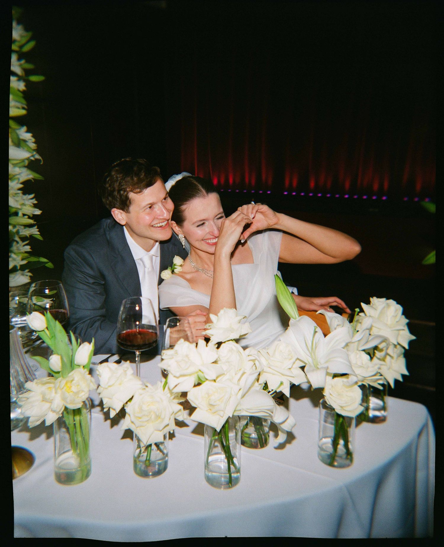 A happy bride and groom sit together at a wedding reception table adorned with white floral arrangements in glass vases.