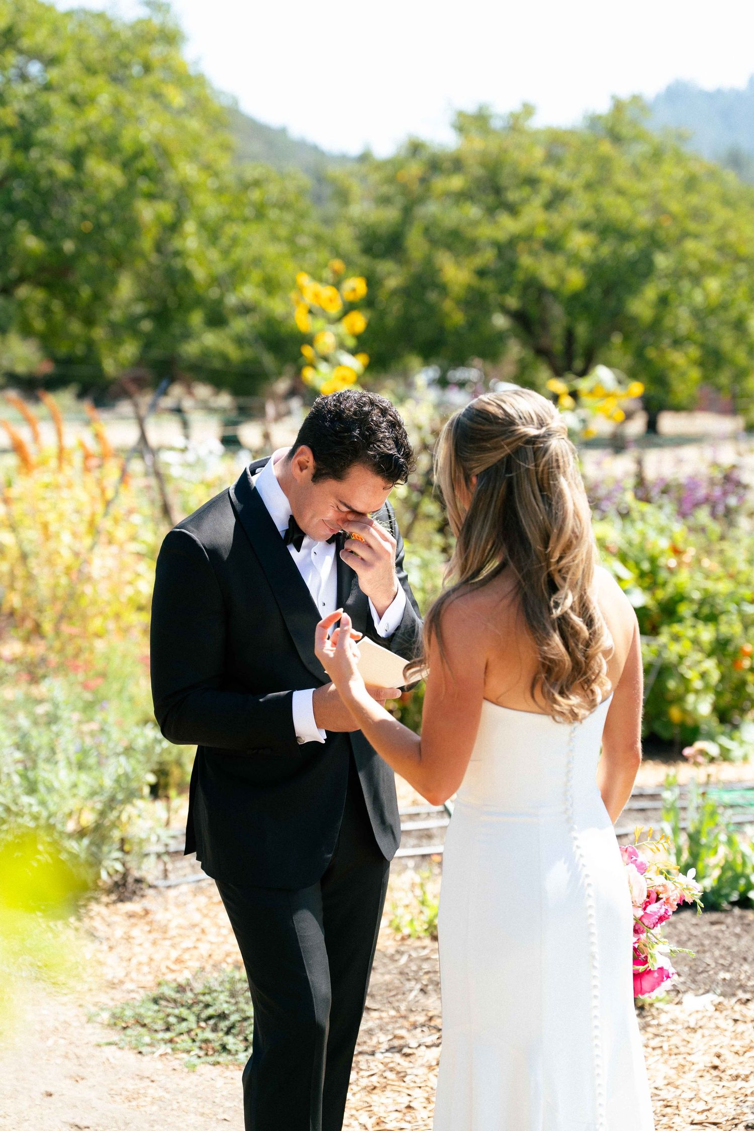 Emotional groom wiping tears during first look moment with bride in sunny garden setting.
