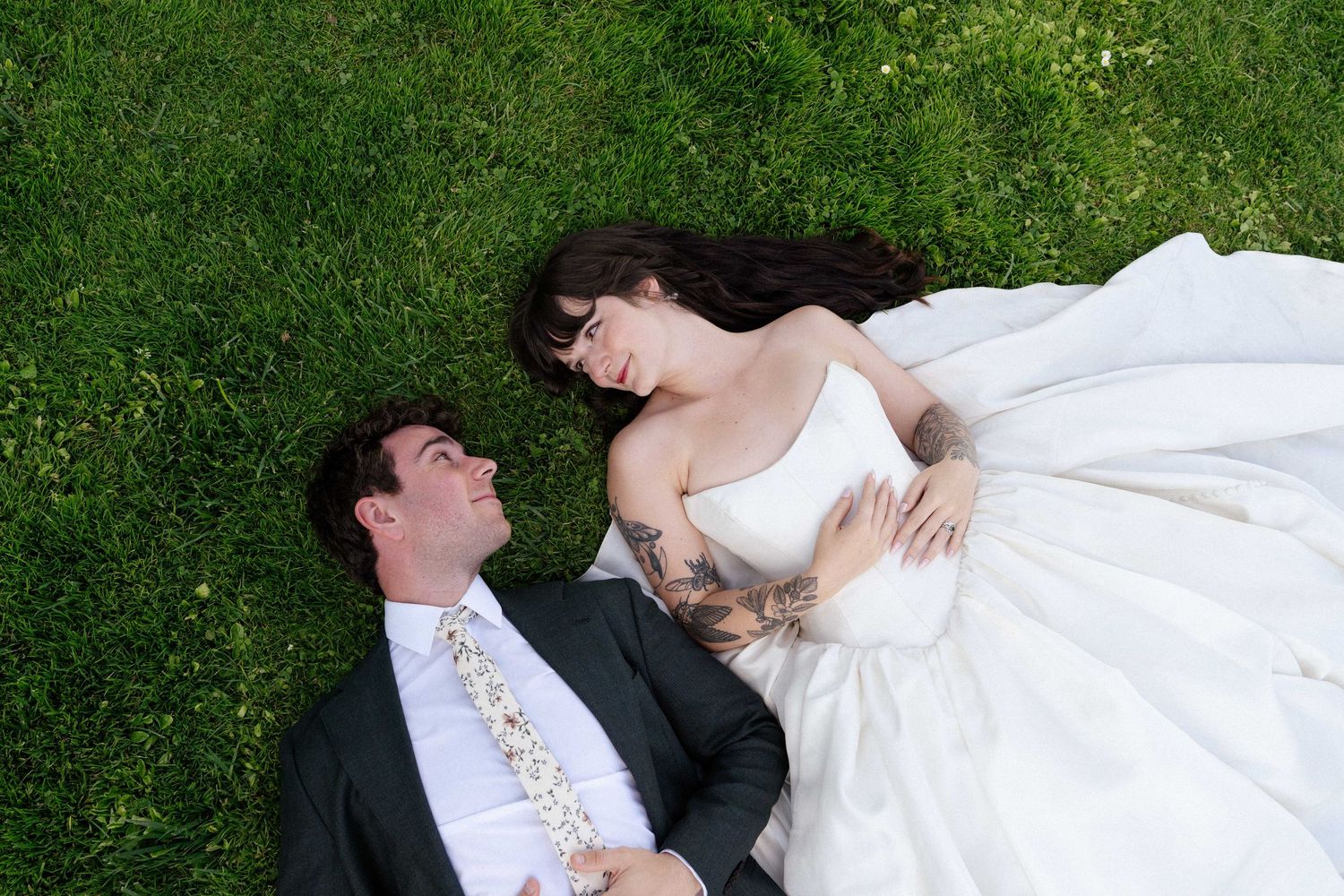 Bride and groom lying on green grass, woman in white wedding dress with tattoos, man in black suit with floral tie.
