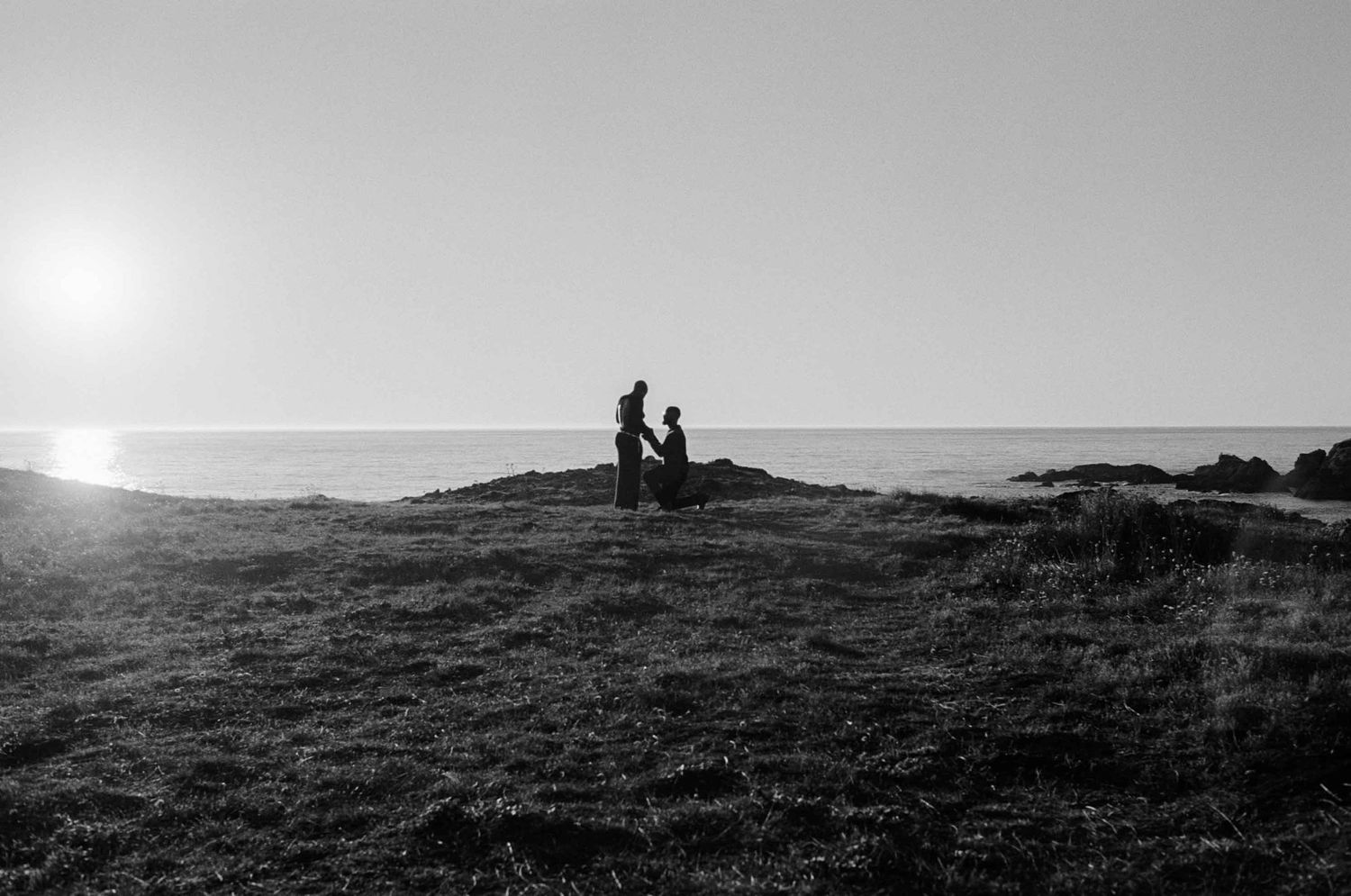 Black and white photo of a couple silhouetted against the ocean horizon at sunset on a grassy cliff.