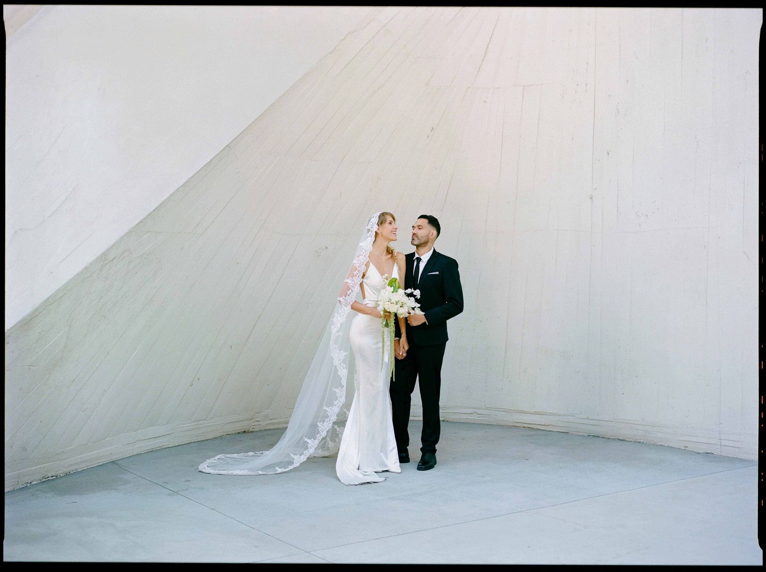 A bride and groom pose elegantly against a minimalist white wall of the TWA Hotel.