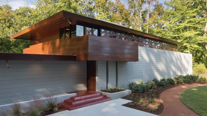 Contemporary mid-century modern home entrance featuring warm wood siding, red steps, and floor-to-ceiling windows amid trees.