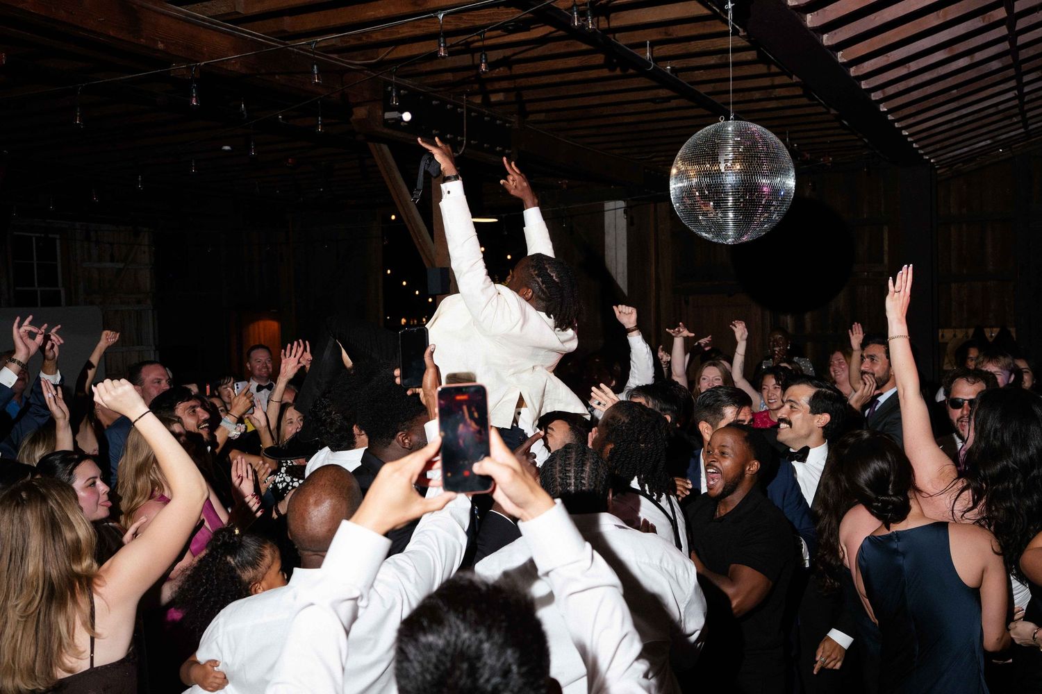 Bride lifted by crowd at lively wedding reception with disco ball and dancing guests.