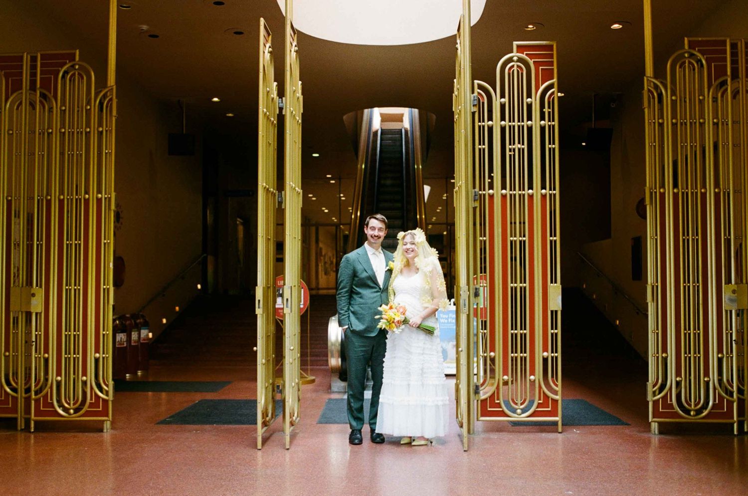 Bride and groom stand smiling in elegant Art Deco gold doorway with escalator backdrop at wedding venue.