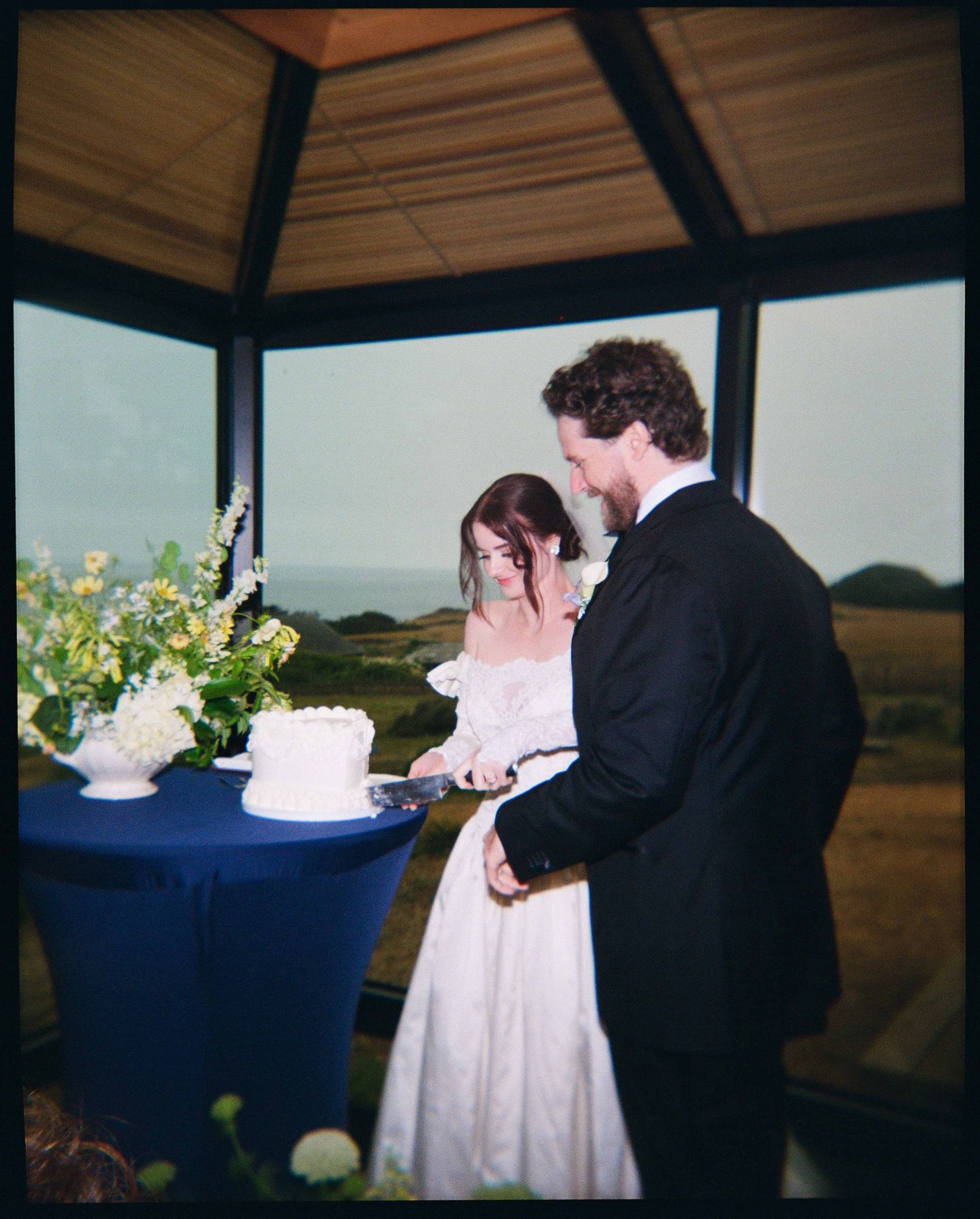 Bride and groom cutting wedding cake under gazebo with ocean view backdrop.